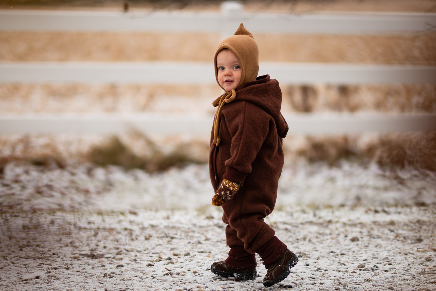Ein Kleinkind in Wollwalk Anzug Kastanie und Zwergenmütze, das im Schnee geht, trägt gestrickte braune und weiße Fäustlinge.