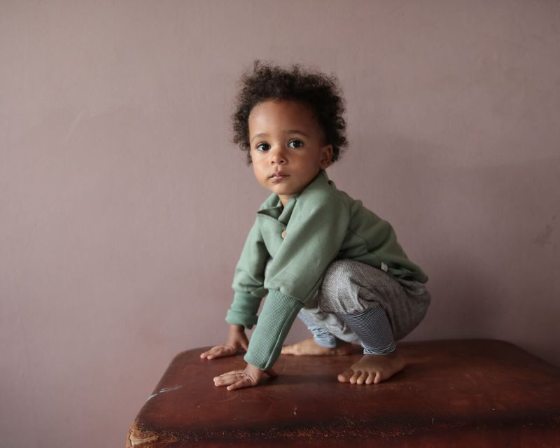 Ein kleiner Junge sitzt auf einem Koffer, seine Füße sind sichtbar. Er trägt ein Schlüttli aus Bio-Baumwolle in Grün.