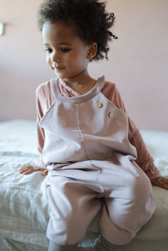 Ein Kind in rosa Latzhose und ein Wickelshirt aus Kaschmir Mix sitzt auf einem Bett. Es handelt sich um ein Kleinkind in einem Innenraum.