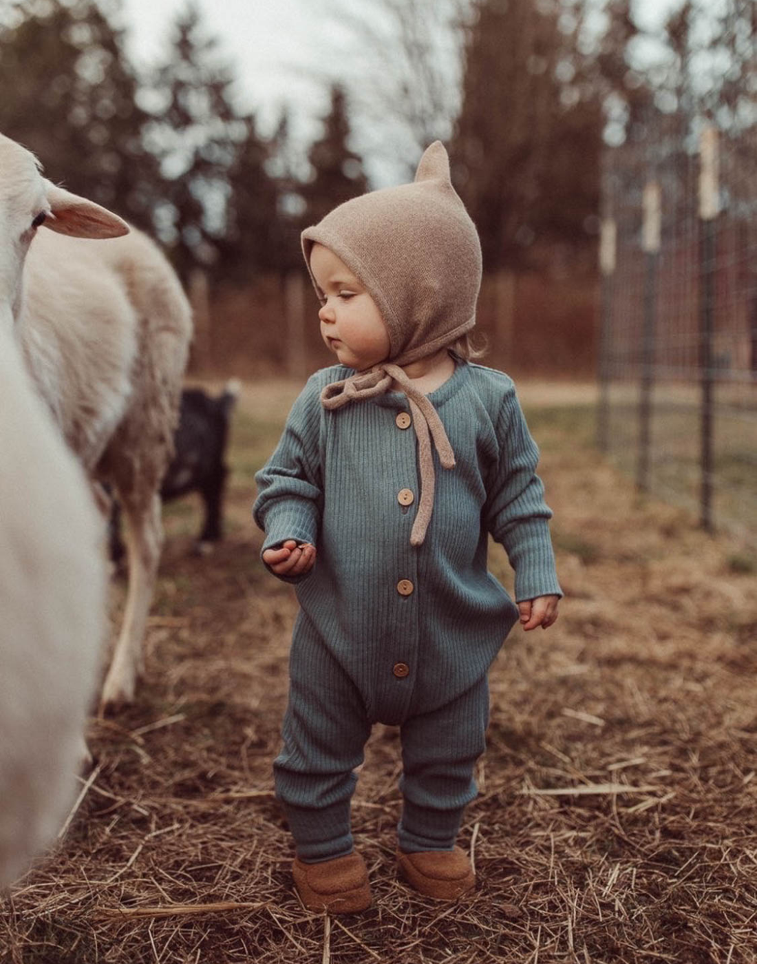Ein Kleinkind in einem eingezäunten Bereich mit Schafen, trägt einen Zwergenmütze Strick und einen Mini Overall Rippstrick, steht auf grünem Gras.