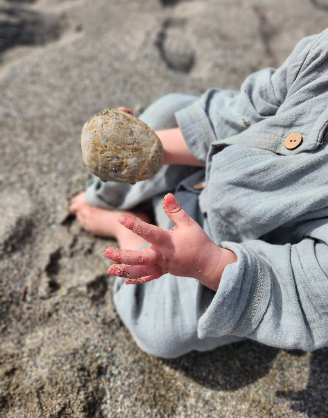 Ein Baby spielt mit Sand am Strand, hat Strampler für Babys für den Sommer aus Musselin an.