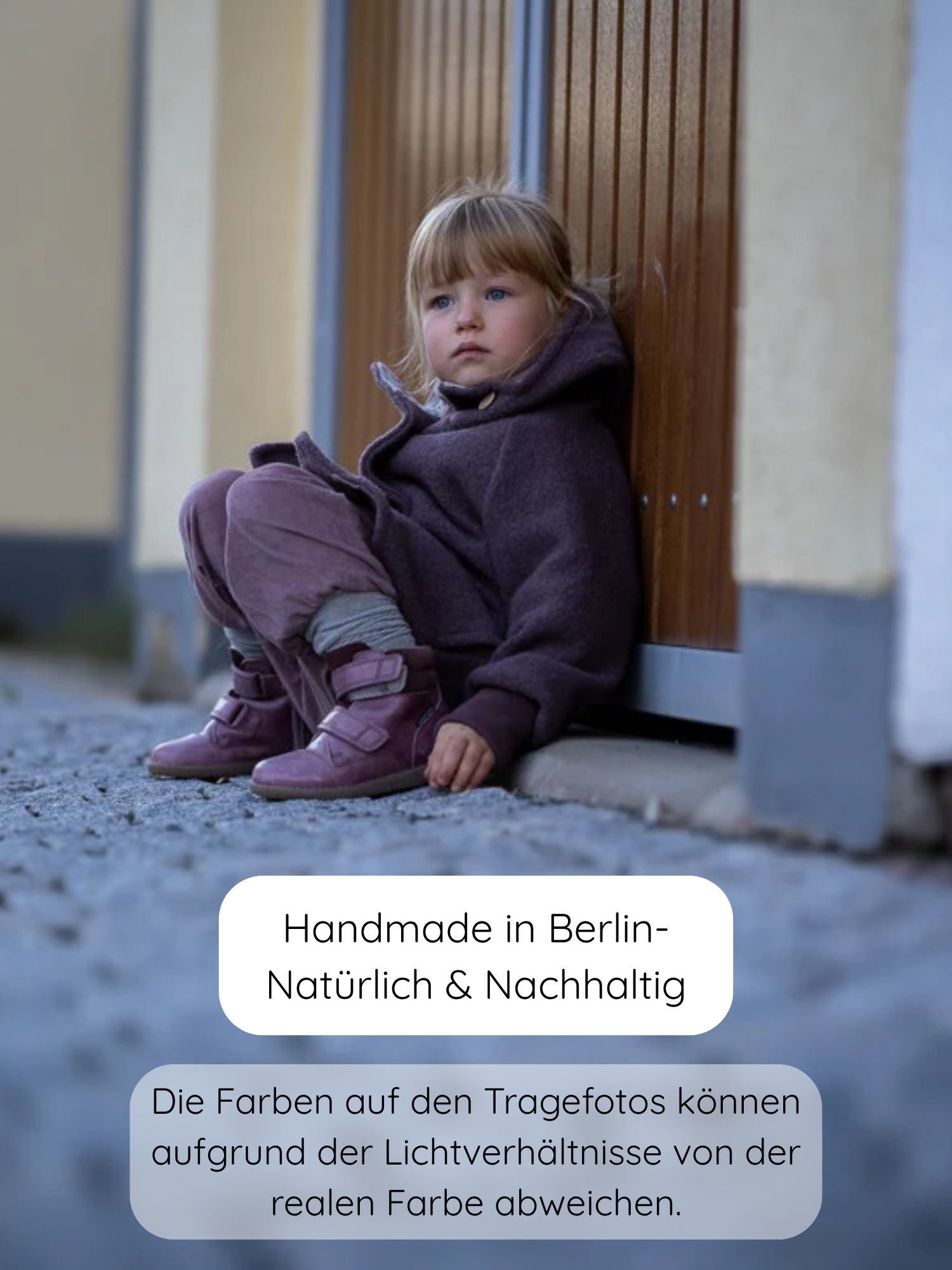 Kleinkind mit blonden Haaren trägt Knickerbocker Feincord Ringel - Malve, bestehend aus einer lila Kapuzenjacke und passender Hose. Es sitzt auf einem Pflasterboden, angelehnt an eine Holzwand.