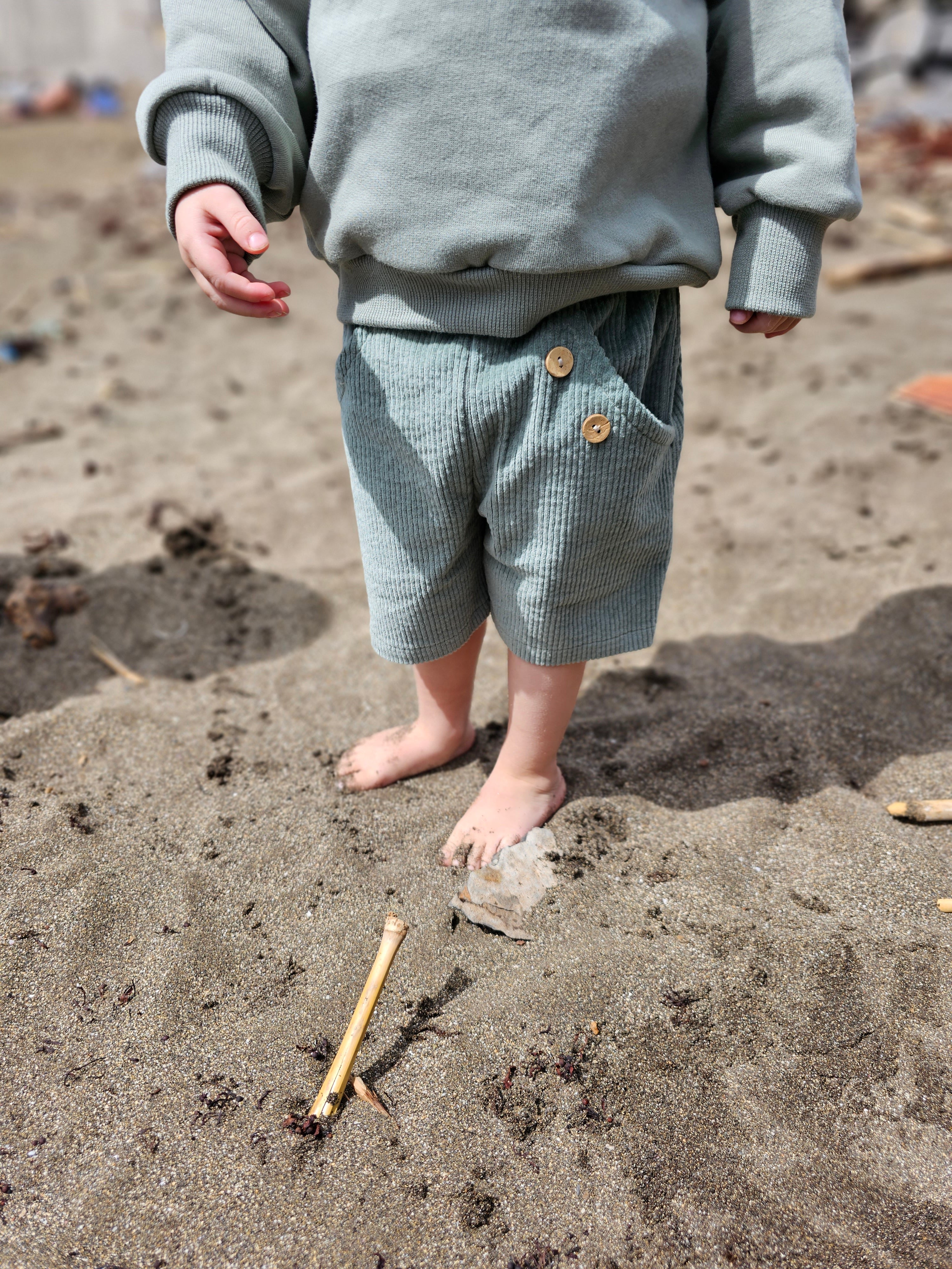 Ein Kind steht barfuß im Sand, trägt die Kurze Buxe Breitcord - Aqua aus weichem Cord, mit Holzknöpfen an der Tasche, umgeben von Treibholz und Seetang.
