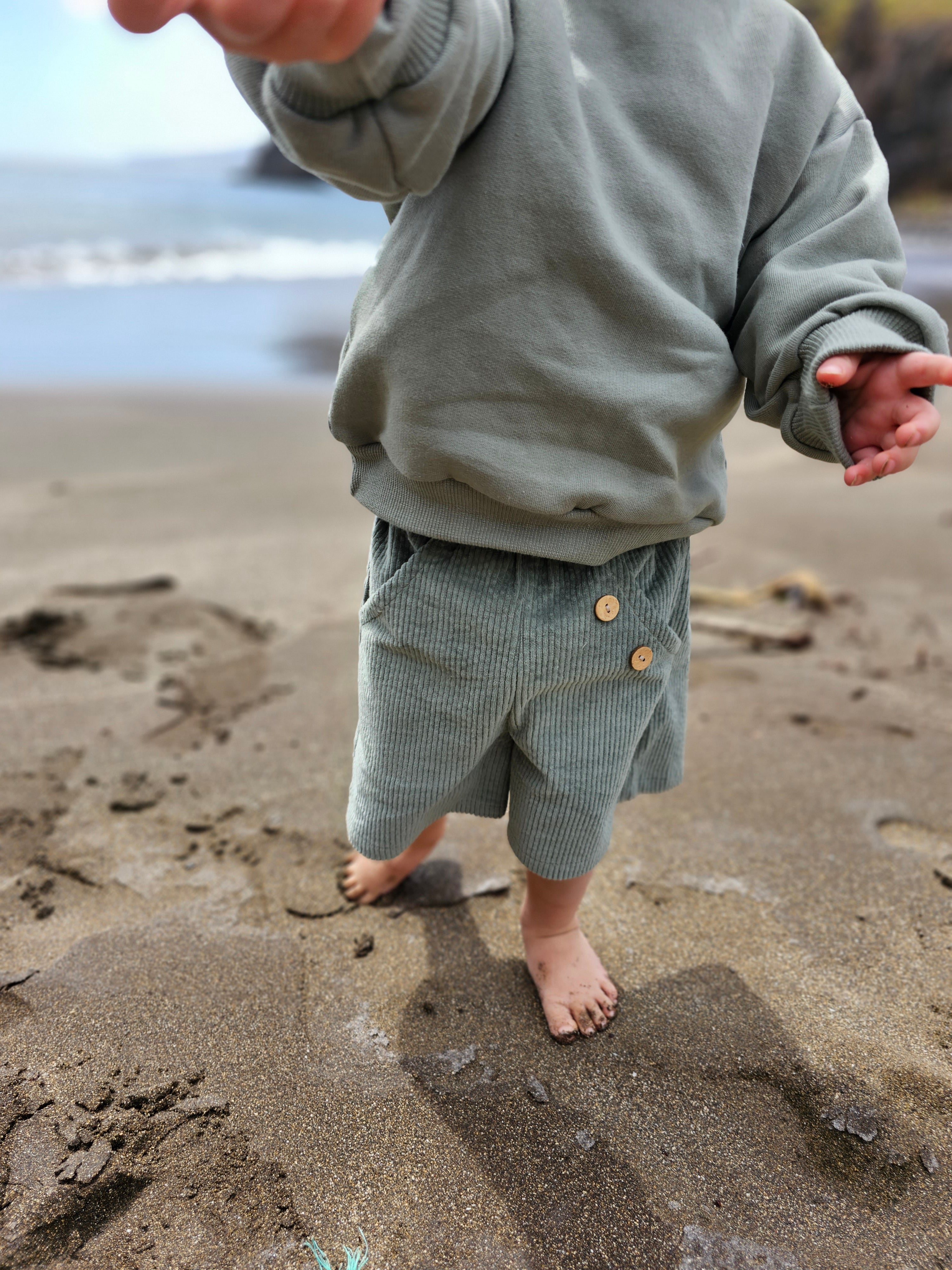Ein Kind trägt die Kurze Buxe Breitcord - Aqua, aus weichem Cord mit Holzknöpfen am Bund, während es barfuß am Strand entlangläuft.