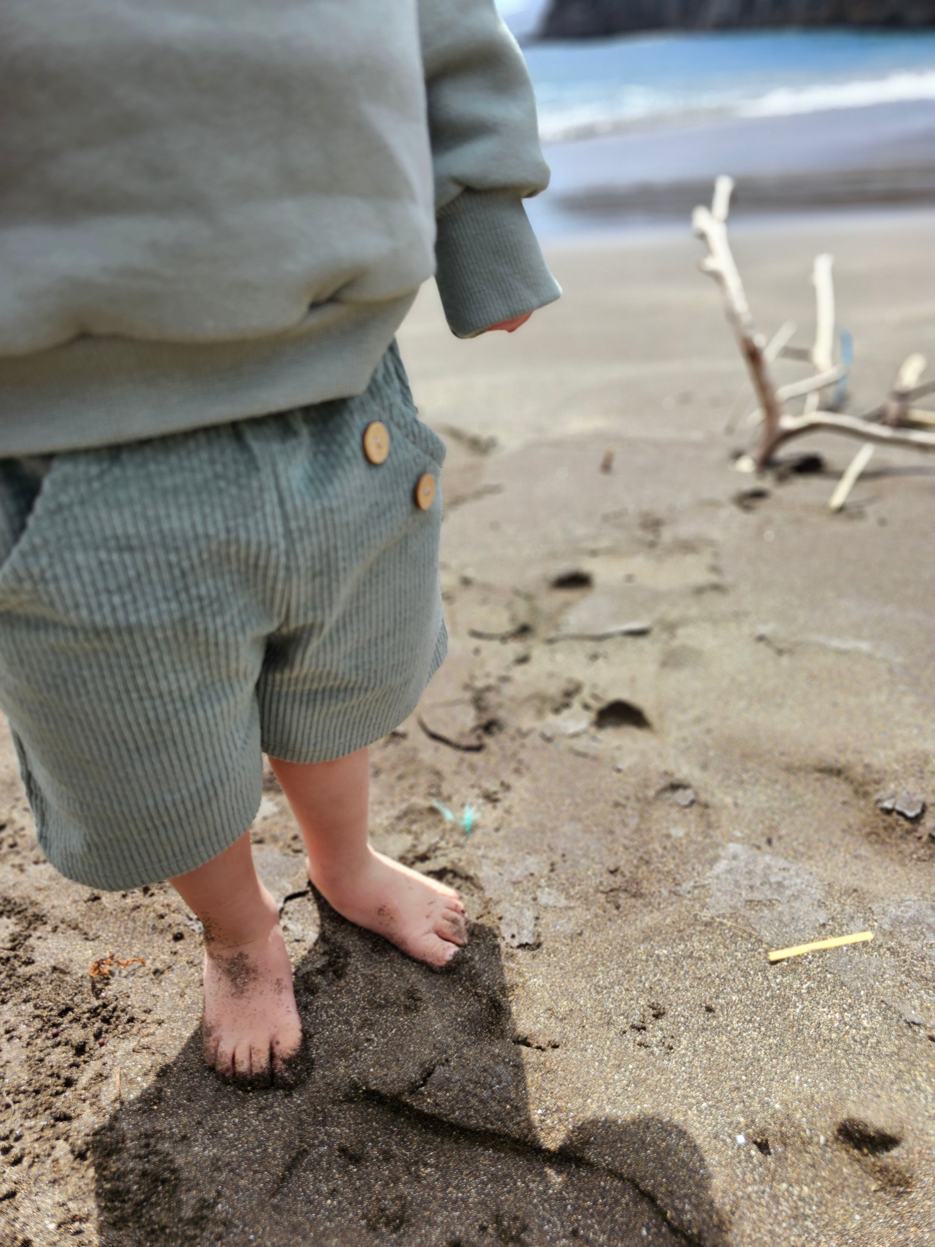 Ein Kind steht barfuß am Strand in sagegrünen Kurze Buxe Breitcord - Aqua Shorts mit Holzknöpfen, umgeben von Sand und Treibholz.