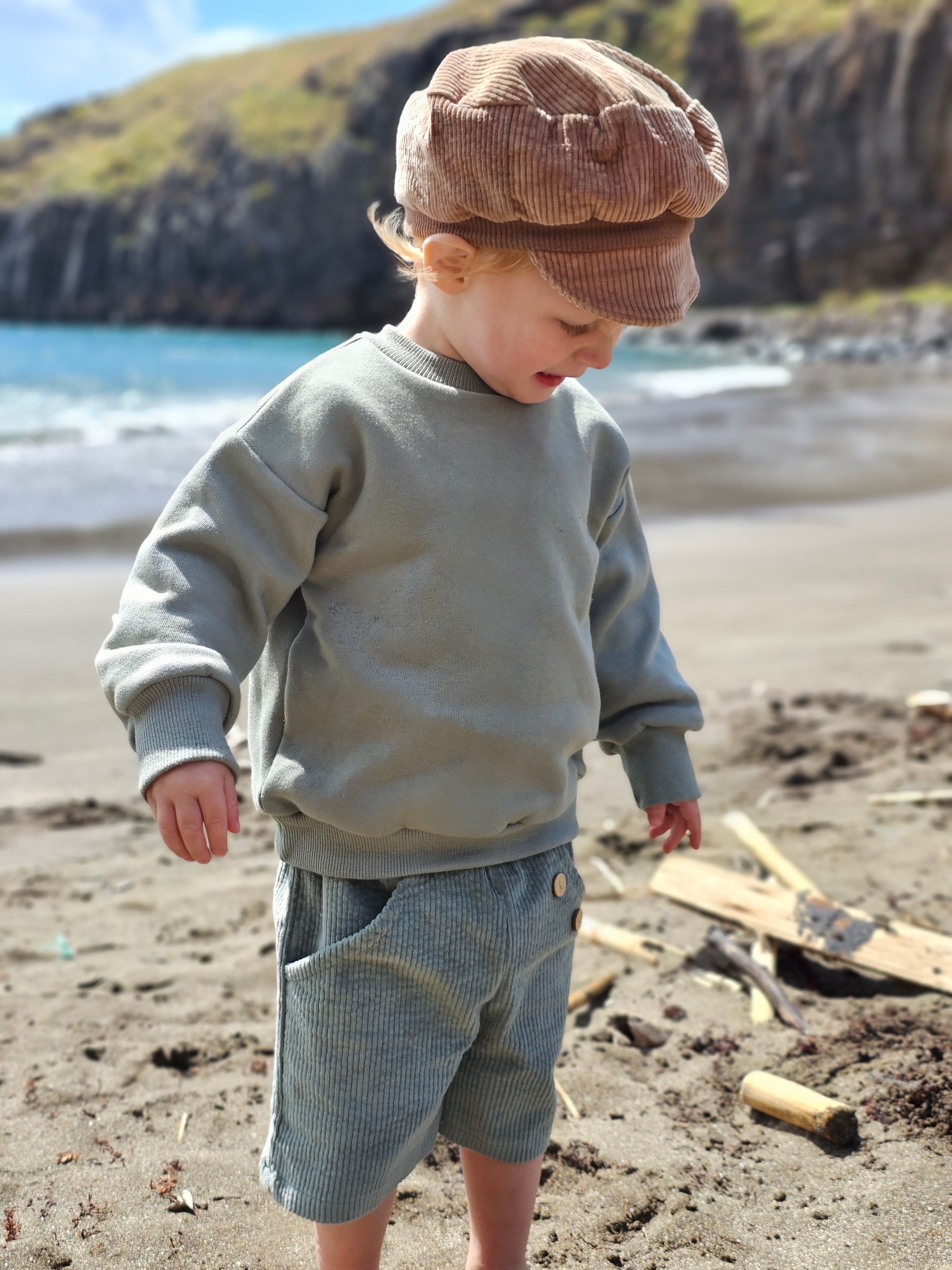 Kleinkind am Strand trägt Kurze Buxe Breitcord - Aqua, eine komfortable, graugrüne Cordhose mit Knopfdetails, ideal für Abenteuer.