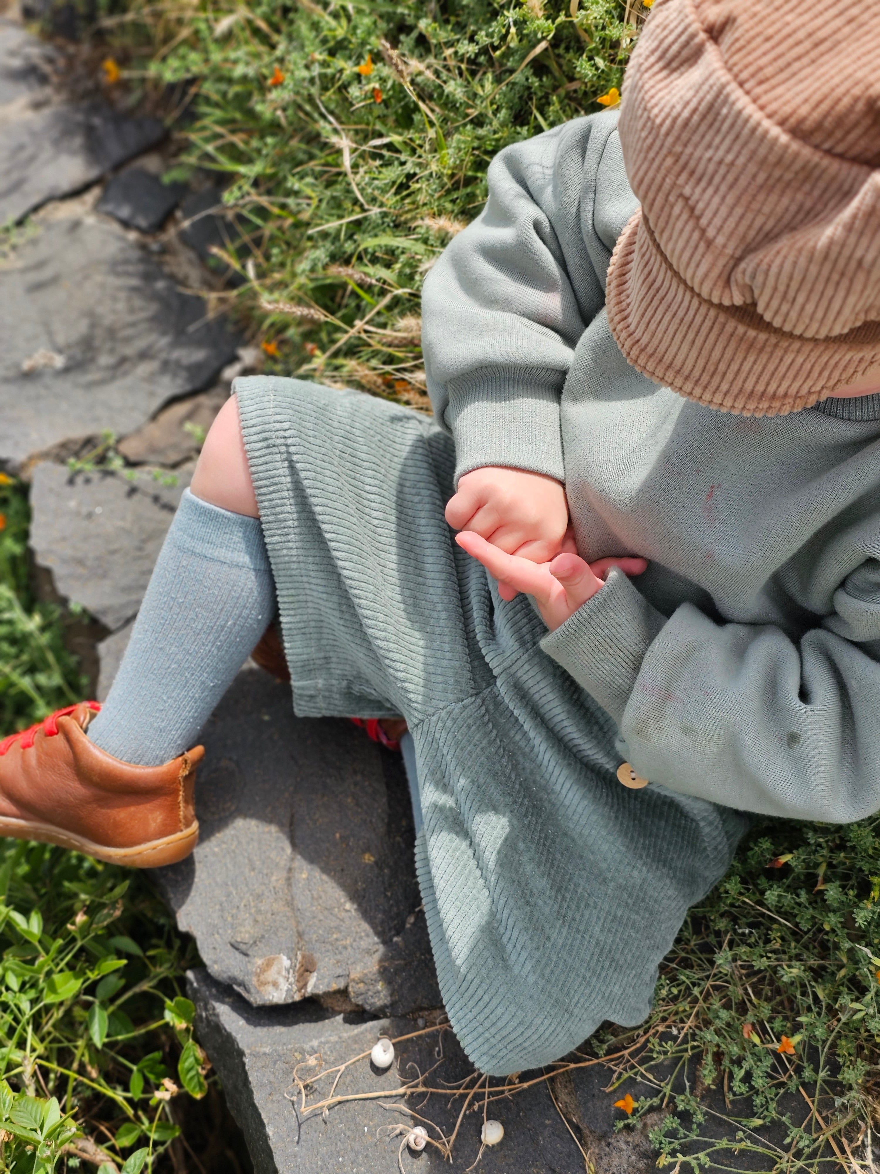 Ein Kind sitzt auf einem Stein, trägt eine Kurze Buxe Breitcord - Aqua mit passenden Kniestrümpfen, braunen Stiefeln und einer Cordmütze, die das Gesicht verdeckt.