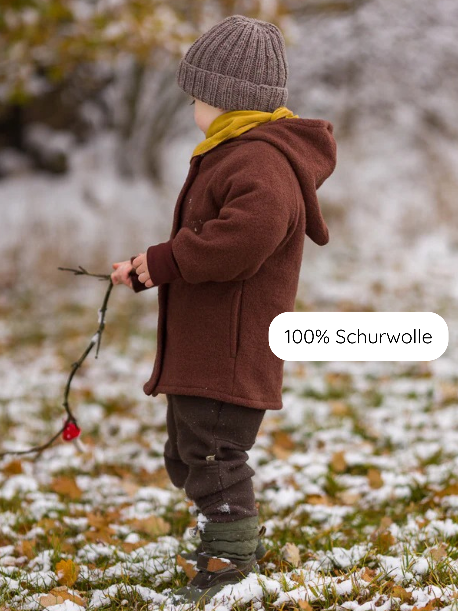 Kleinkind im Profil, trägt Winter Wichteljacke Wollwalk - Schoko mit spitzer Kapuze, kombiniert mit einem gelben Schal und dunklen Wollhosen. Es hält einen Zweig mit roter Beere.