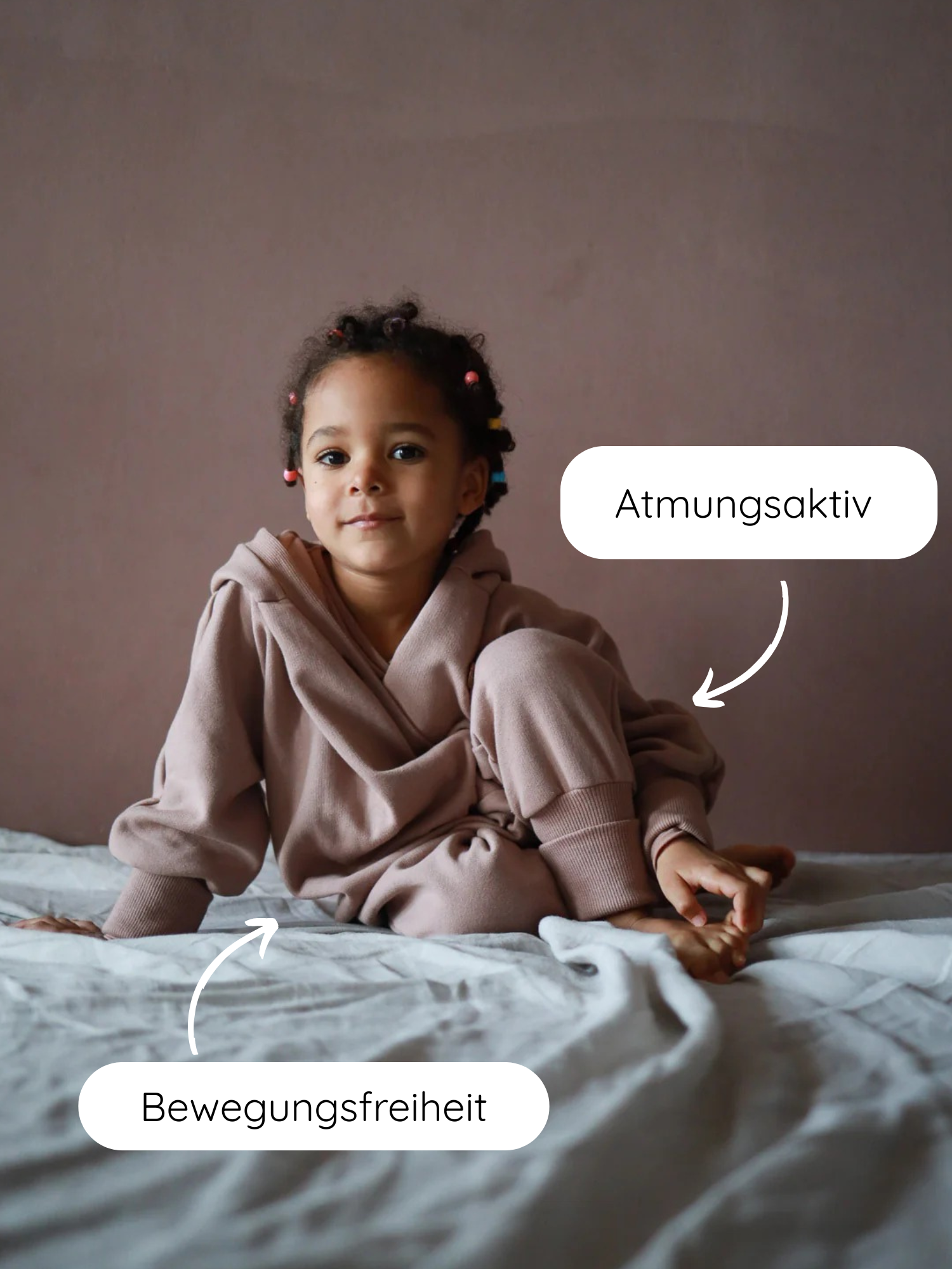 Ein Kind trägt den Kuschelanzug Bio-Sweat - Puder, sitzt barfuß auf einem Bett. Call-outs zeigen Bewegungsfreiheit an der Hüfte und Atmungsaktiv am Knie.