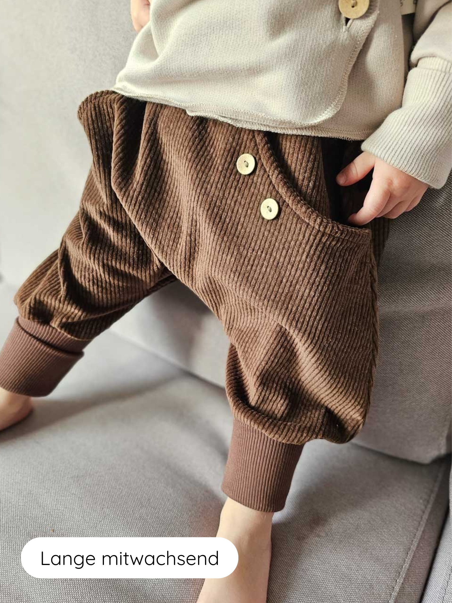 Knickerbocker Breitcord - Schoko: Ein Kind trägt locker sitzende, schokoladenbraune Cordhosen mit elastischem Bund, Holzknöpfen und gerippten Bündchen, auf einer weichen, hellgrauen Oberfläche sitzend.