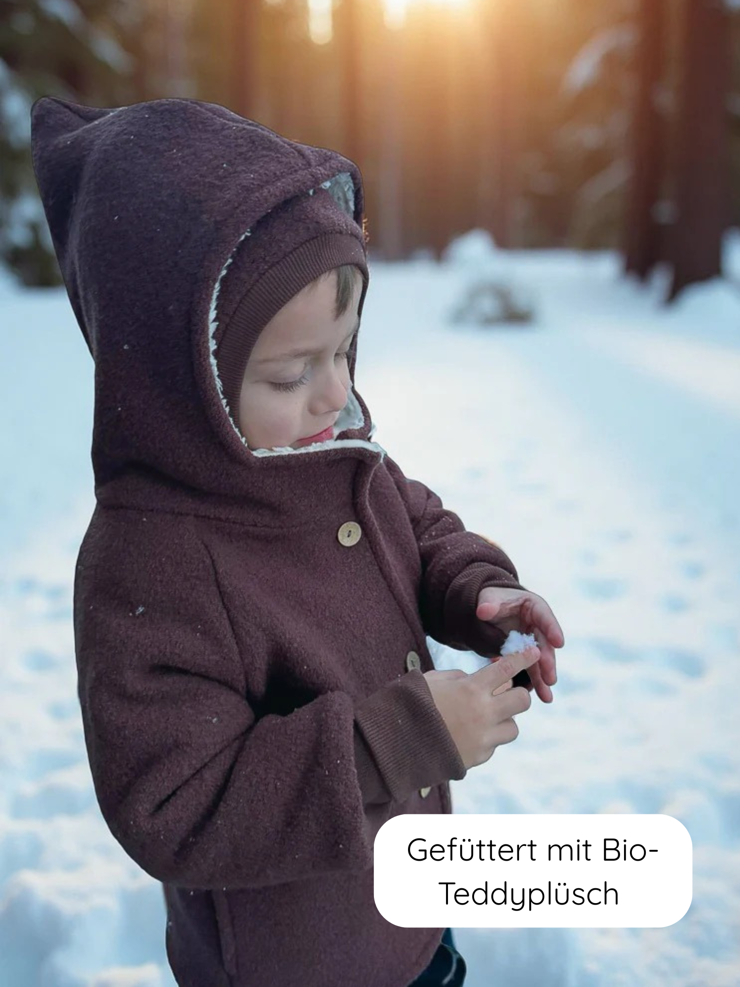 Kleinkind im Wald trägt Winter Wichteljacke Wollwalk - Schoko, mit spitzer Kapuze und breiten Bündchen, untersucht Schnee bei Sonnenuntergang. Text: „Gefüttert mit Bio-Teddyplüsch“.
