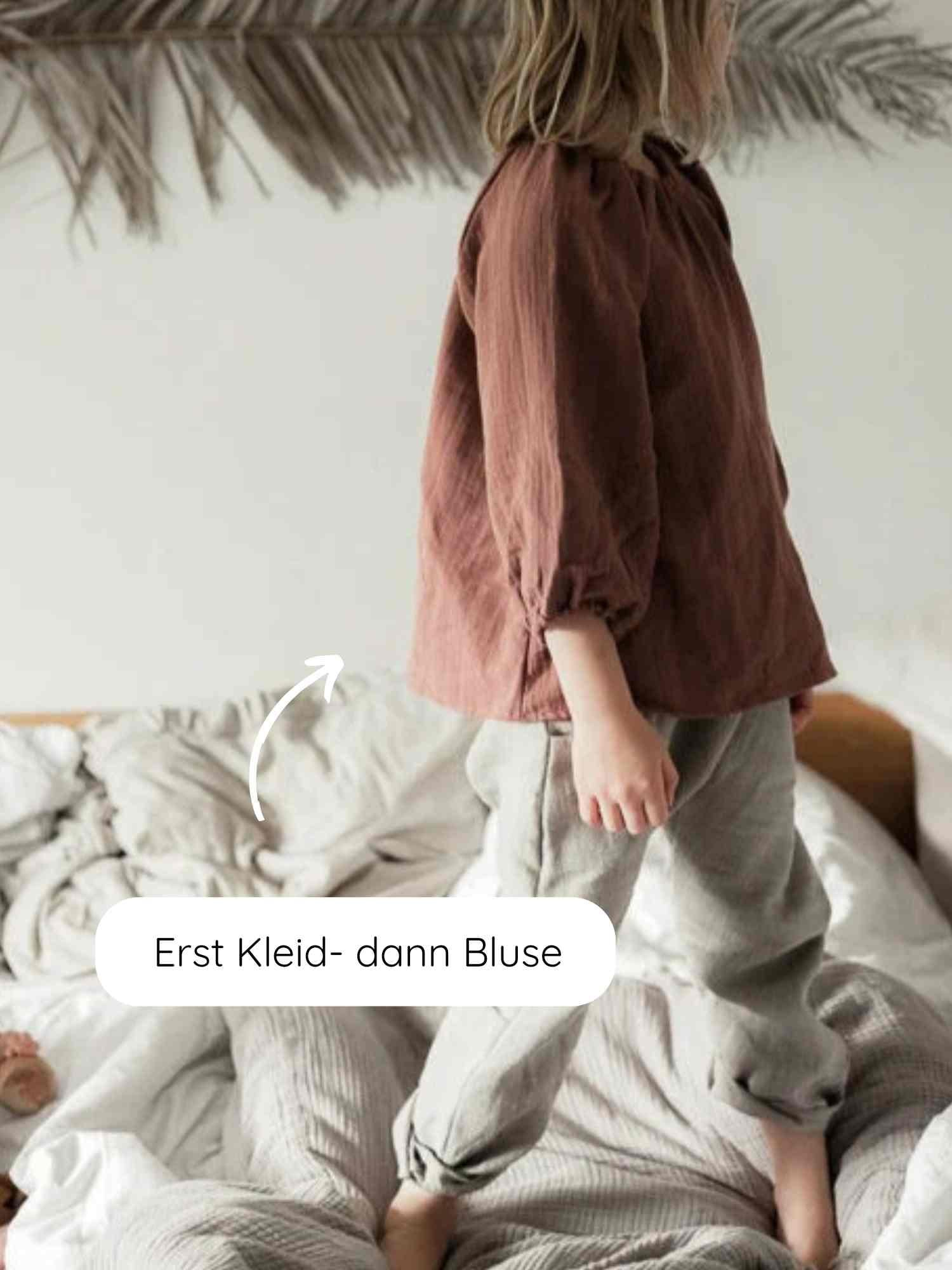 Kind im Profil auf ungemachtem Bett trägt Blüschen Musselin - Malve, locker geschnitten, ideal für warme Tage. Sprechblase zeigt: Erst Kleid – dann Bluse. Canopy aus Palmblättern im Hintergrund.