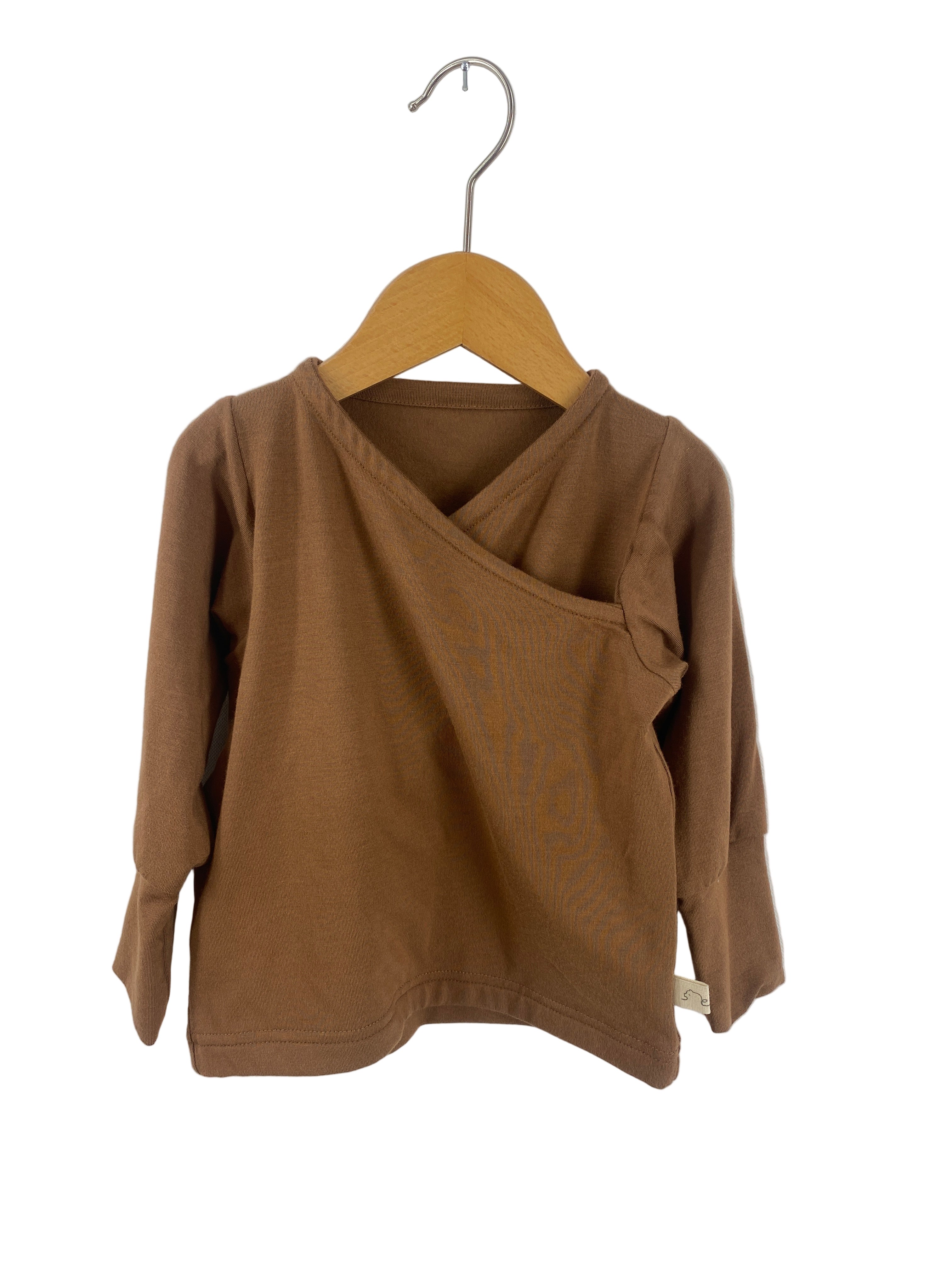 Wickelshirt Tencel Muskat Gr 1Y: Ein kleines, langärmeliges Oberteil mit Wickelausschnitt, hängend an einem einfachen Holzkleiderbügel. Neuware mit kleinem Stoffetikett an der unteren Seitennaht.