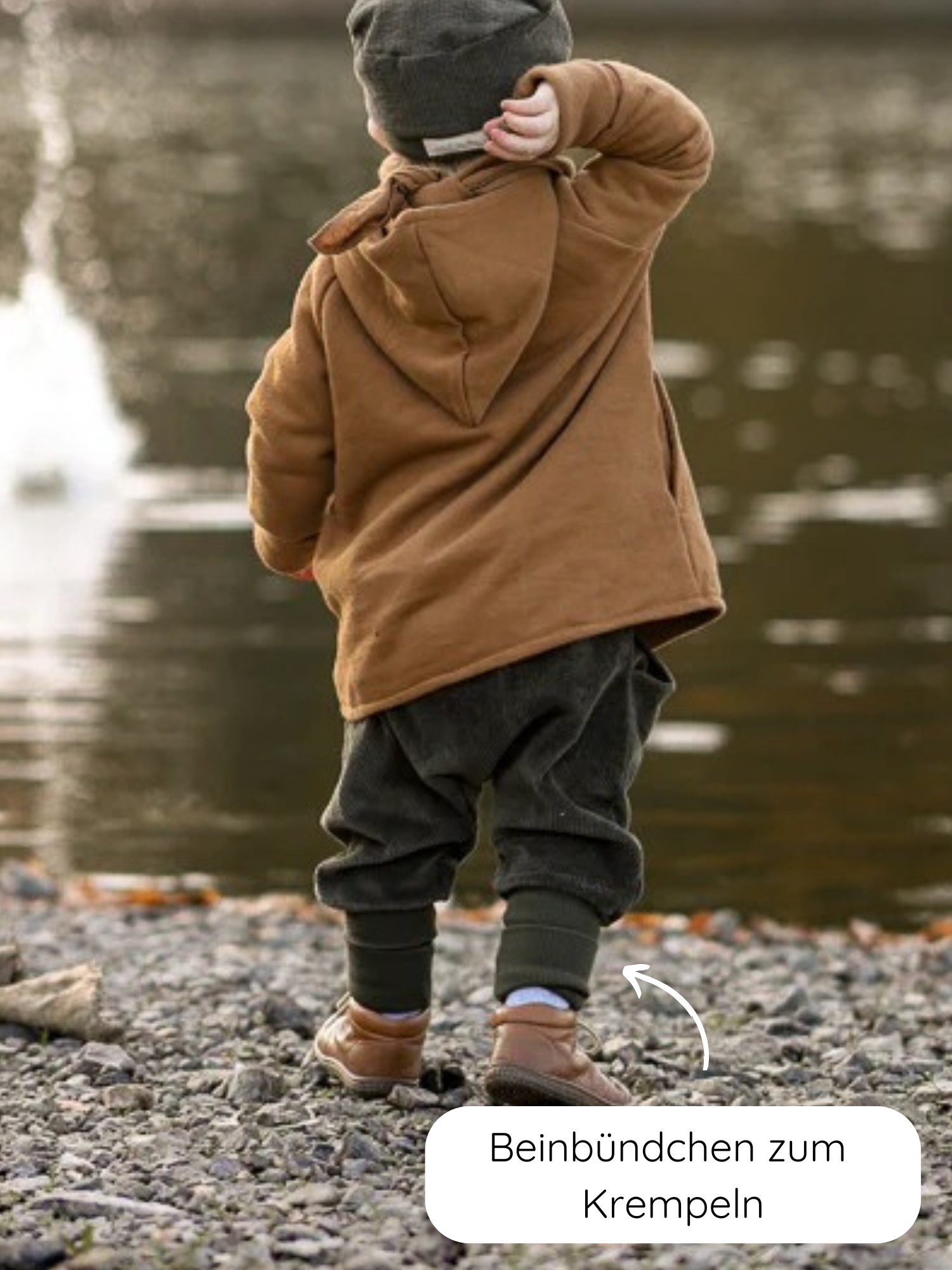 Ein Kind am Seeufer trägt die Knickerbocker Breitcord - Kiwi mit aufrollbaren Beinbündchen, kombiniert mit einer braunen Kapuzenjacke, dunklen Hosen, einer grauen Mütze und braunen Schuhen.