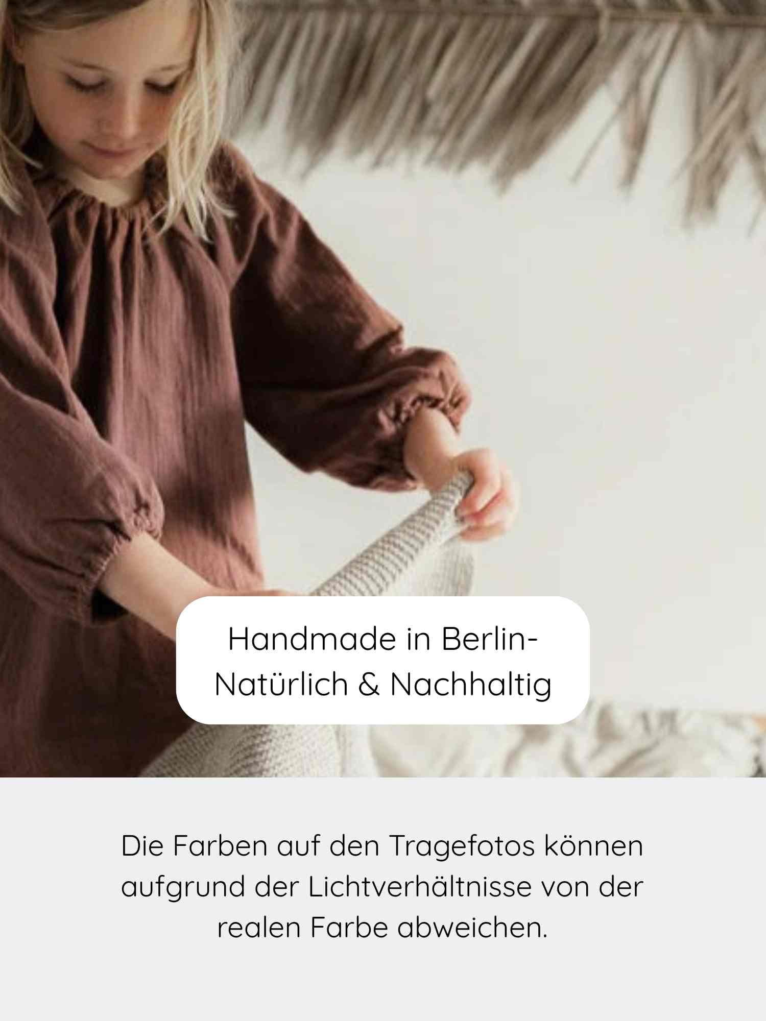 Mädchen in lockerem, langärmligem Kleid hält handgestrickte Decke. Unten Text Handmade in Berlin. Produkt: Blüschen Musselin - Malve, ideal für empfindliche Kinderhaut, locker und atmungsaktiv.