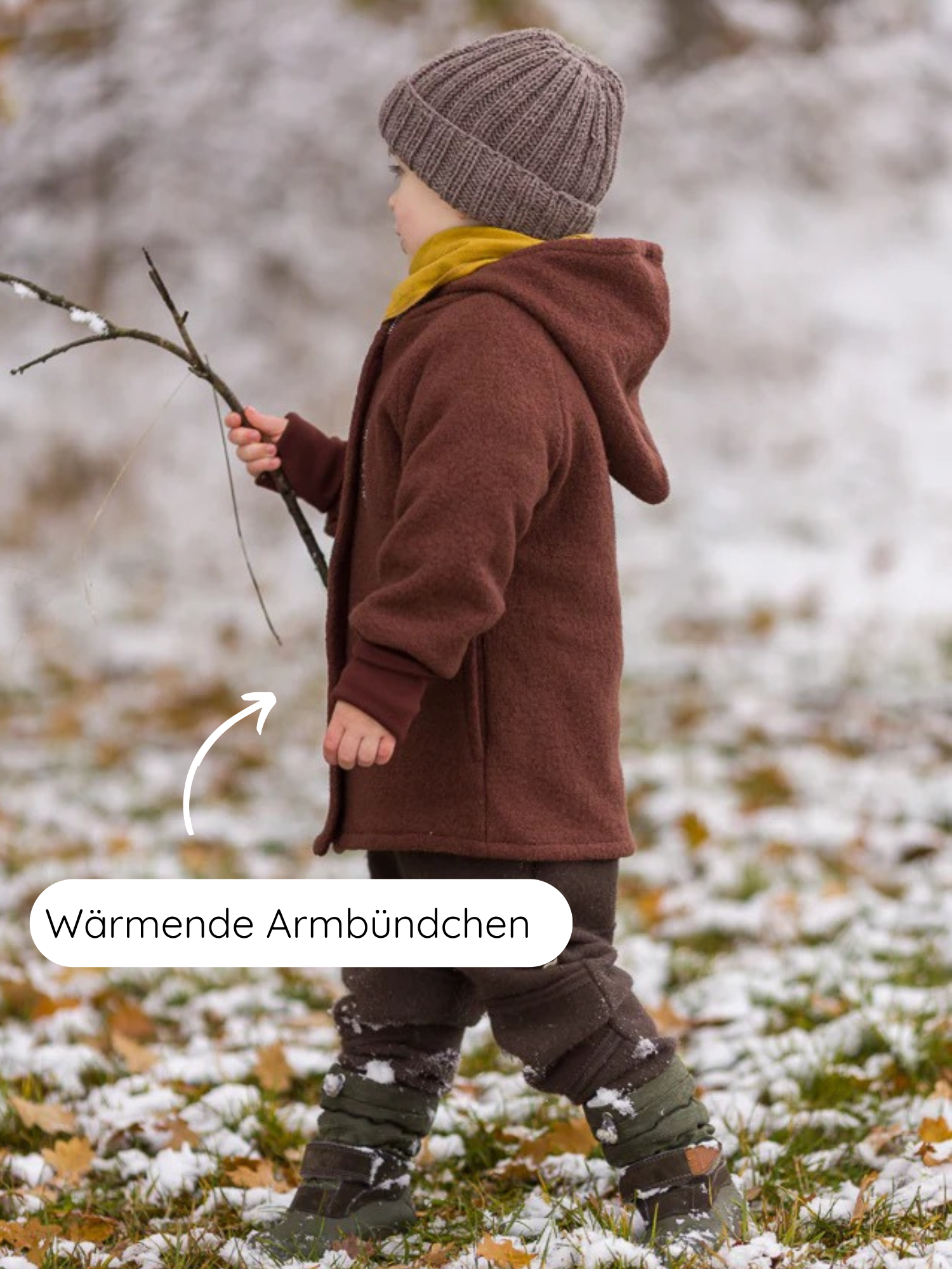 Kleinkind im Freien, trägt eine Winter Wichteljacke Wollwalk - Schoko mit spitzer Kapuze und wärmenden Armbündchen, dazu Mütze, dunkle Hosen und warme Stiefel, hält einen Ast.
