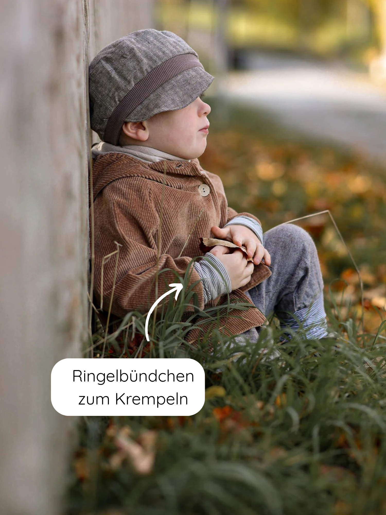 Kleinkind in Wichteljacke Breitcord Stretch - Weizen mit Ringel-Armbündchen sitzt auf Gras, trägt graue Hose und Kappe, hält Herbstblatt. Jacke zeigt aufrollbare Streifenbündchen, ideal für Übergangszeit.