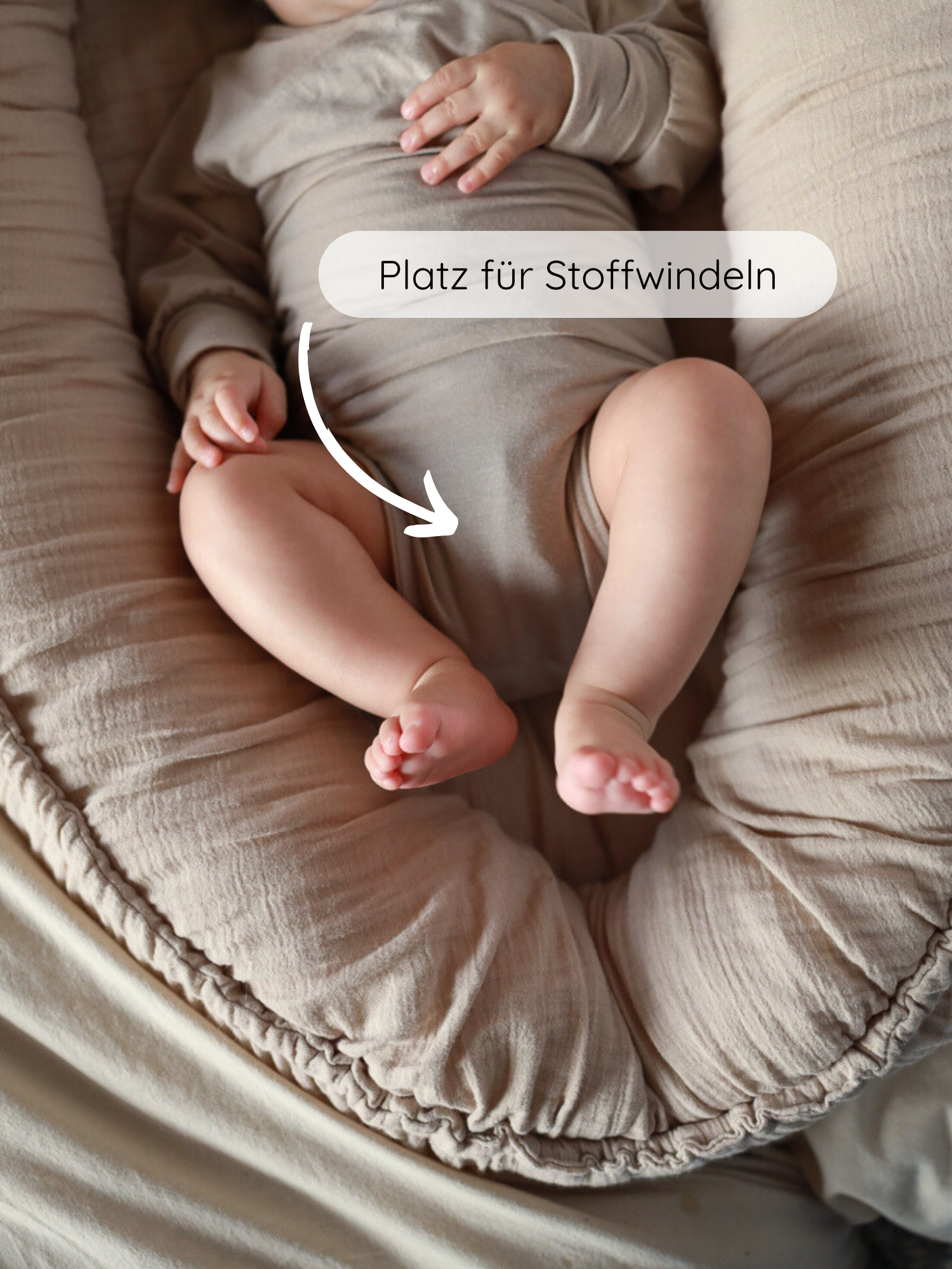 Baby in einem Loopbody Kaschmir-Mix - Sand, liegend in einem gepolsterten Babynest. Ein Pfeil zeigt auf den Schrittbereich, beschriftet mit “Platz für Stoffwindeln”, betont die Bequemlichkeit und Passform.