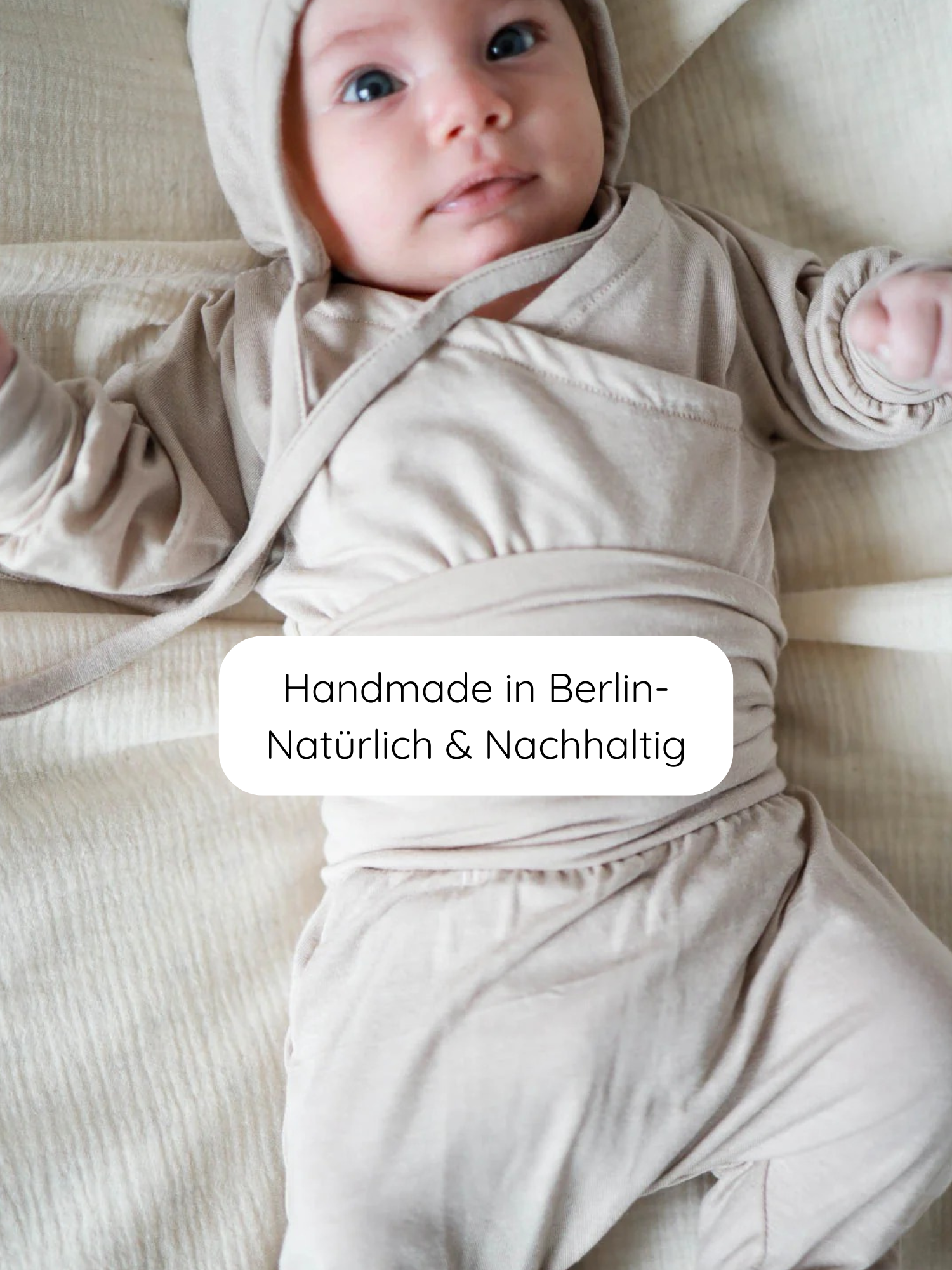 Kleinkind in Loopbody Kaschmir-Mix - Sand, weiches Wickel-Set mit Kapuze und Hose, liegt auf passender Decke. Label: Handmade in Berlin – Natürlich & Nachhaltig sichtbar auf dem Bauch.