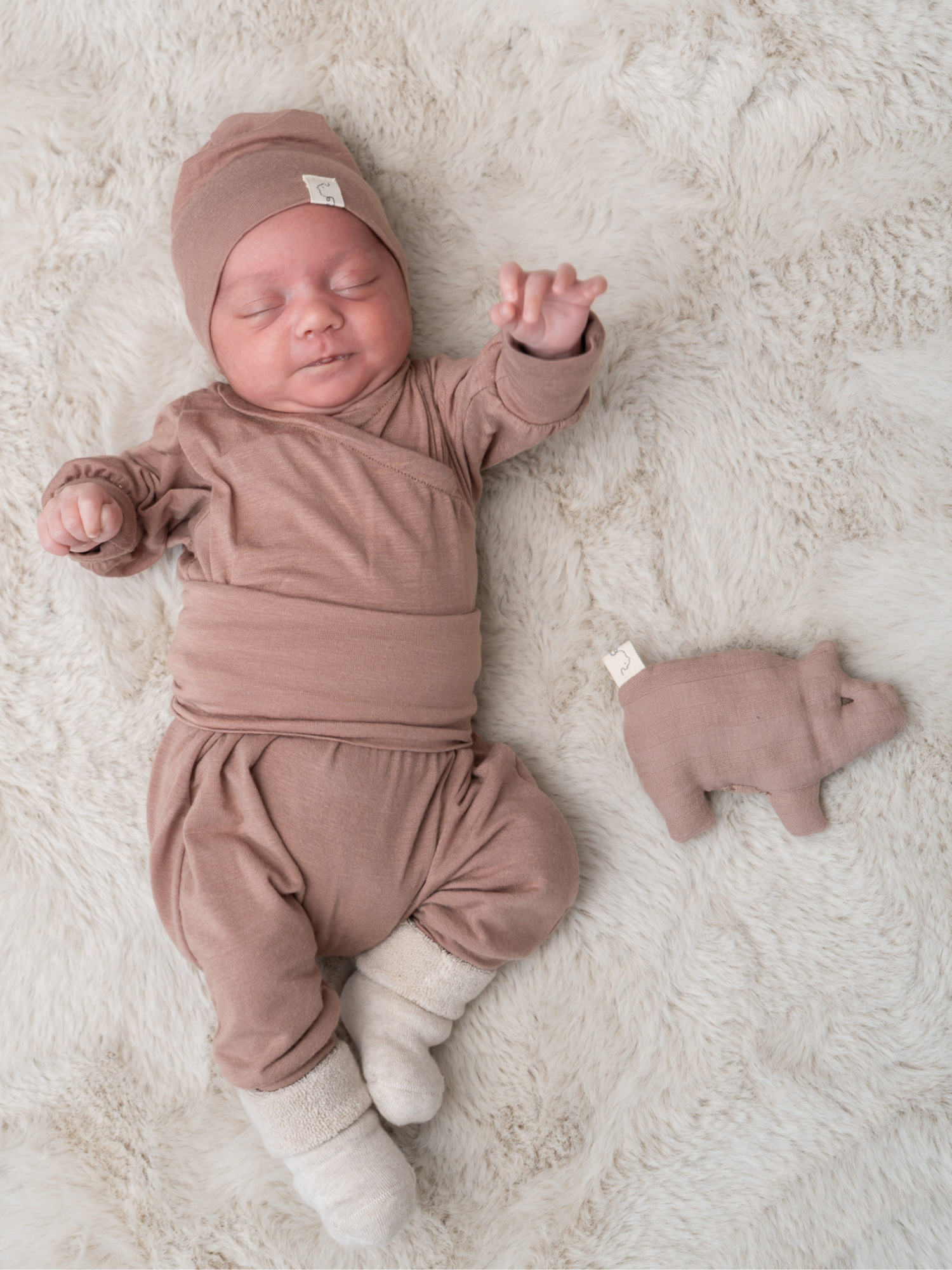 Babyknicker Kaschmir-Mix - Puder: Neugeborenes in weichem, taupefarbenem Strickoutfit mit passendem Mützchen und cremefarbenen Socken, neben einem passenden Strick-Plüschschweinchen auf einer Kunstfell-Decke.