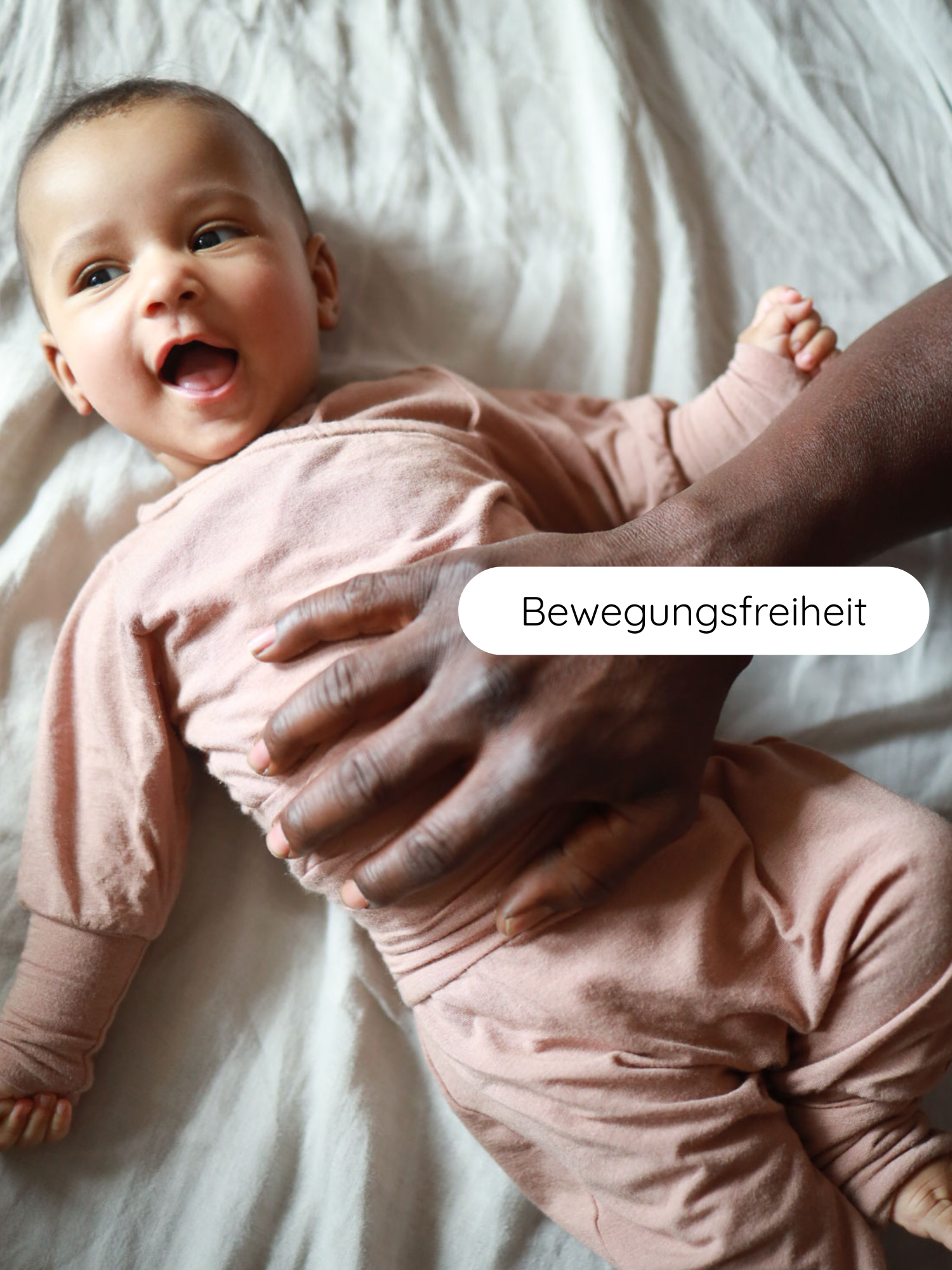 Babyknicker Kaschmir-Mix - Puder: Ein lächelndes Baby in einem weichen, rosa Outfit liegt auf einem hellen Laken, gehalten von dunkleren Händen. Text Bewegungsfreiheit betont die bequeme Passform.