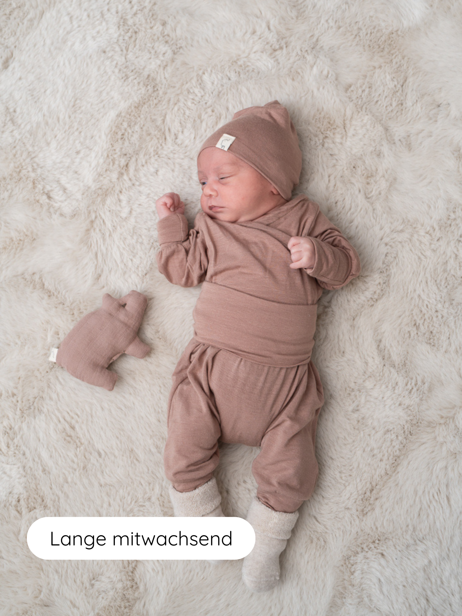 Neugeborenes auf einer flauschigen Decke in Babyknicker Kaschmir-Mix - Puder mit passendem Hut, Oberteil, und Stiefelchen, neben einem Stofftier; Text Lange mitwachsend zeigt, dass die Kleidung mitwächst.