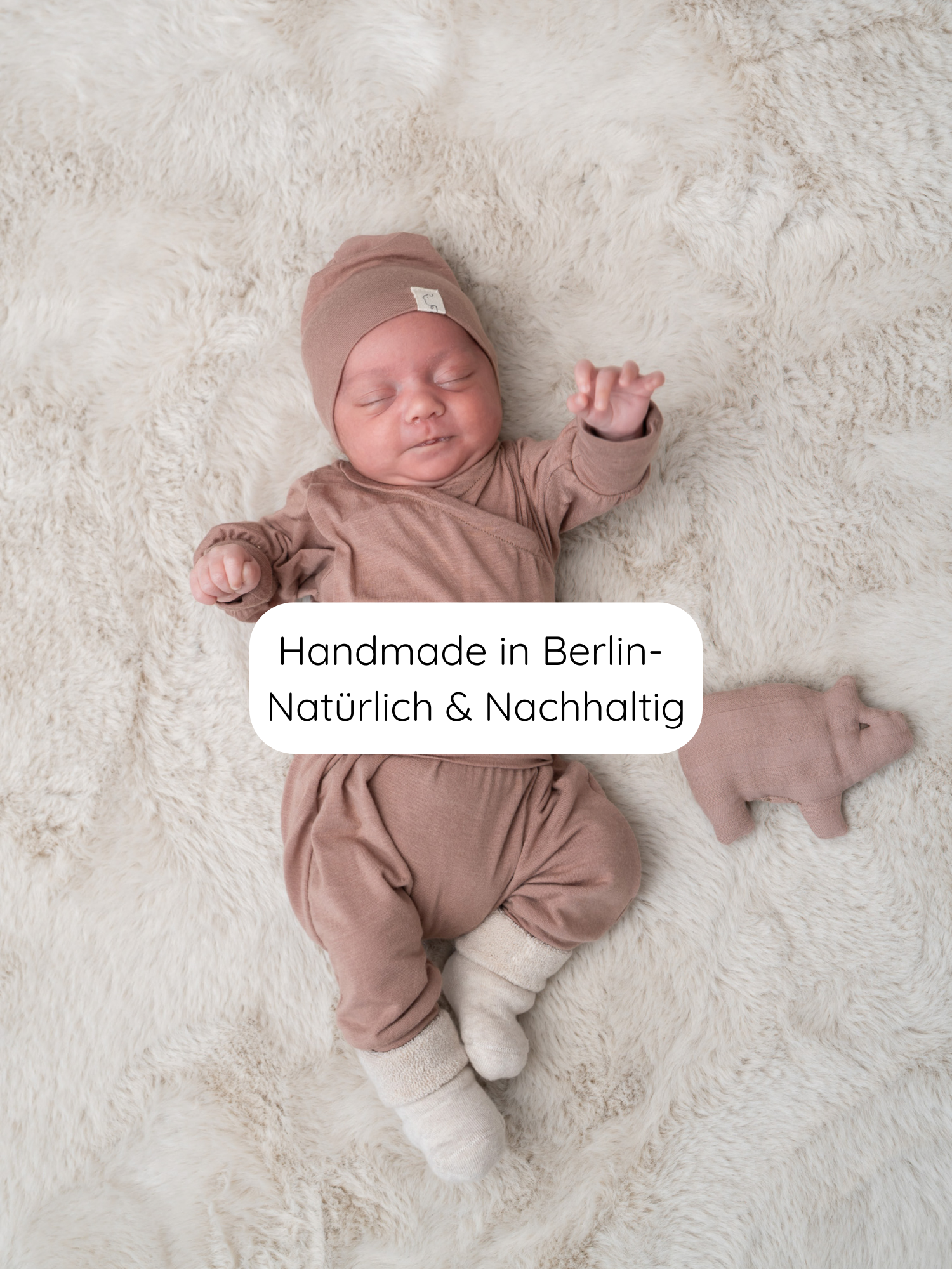 Neugeborenes auf weicher Decke trägt Babyknicker Kaschmir-Mix - Puder mit Klappbündchen, passendem Mützchen und Socken. Daneben ein kleines Stofftier. Text: “Handmade in Berlin – Natürlich & Nachhaltig”.