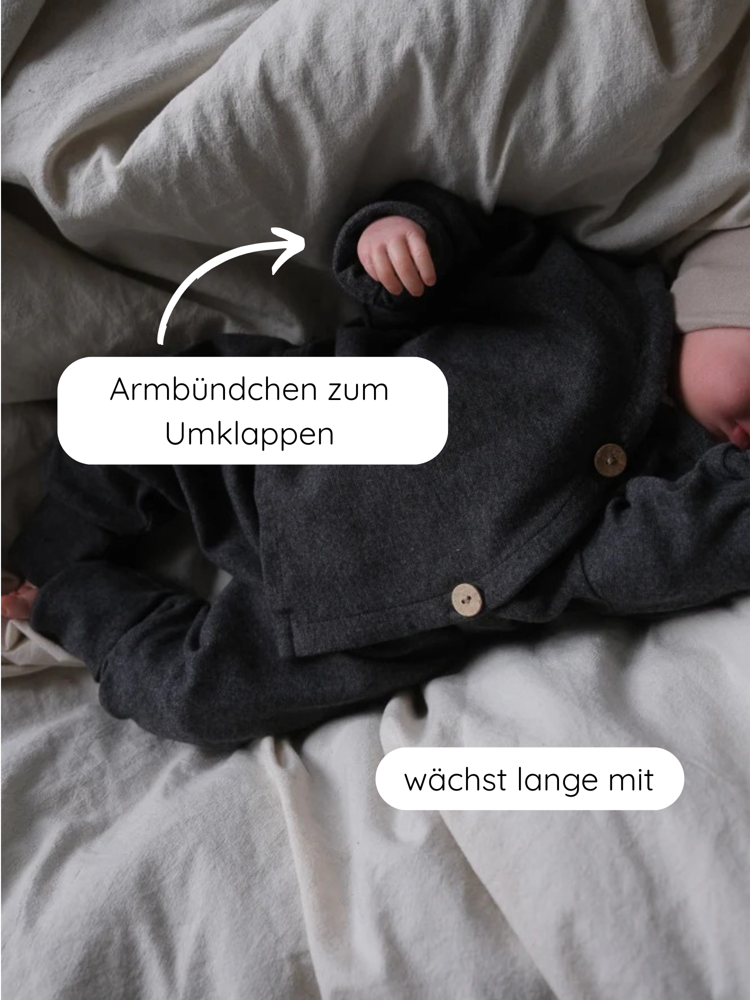 Babyschlüttli Feinstrick - Ruß Meliert: Ein Baby liegt in einem dunklen, melierten Strick-Romper mit umklappbaren Armbündchen und mitwachsendem Design auf dem Bett. Ideal für Komfort und Stil.