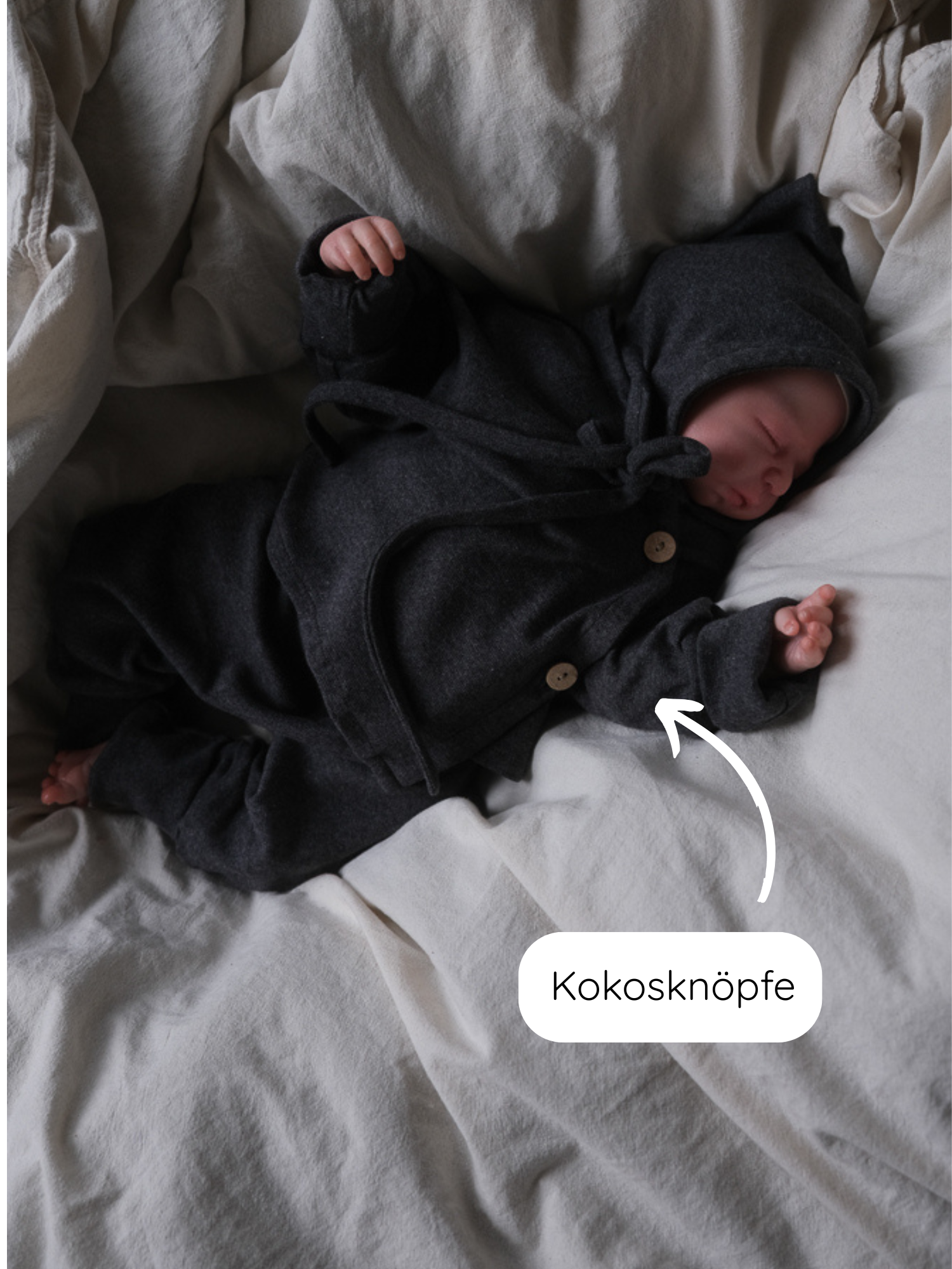 Neugeborenes in einem Babyschlüttli Feinstrick - Ruß Meliert aus Baumwolle-Elasthan-Mix mit Kokosknöpfen, liegt auf cremefarbener Bettwäsche. Wickelverschluss und Kapuze bieten Funktionalität und Komfort.