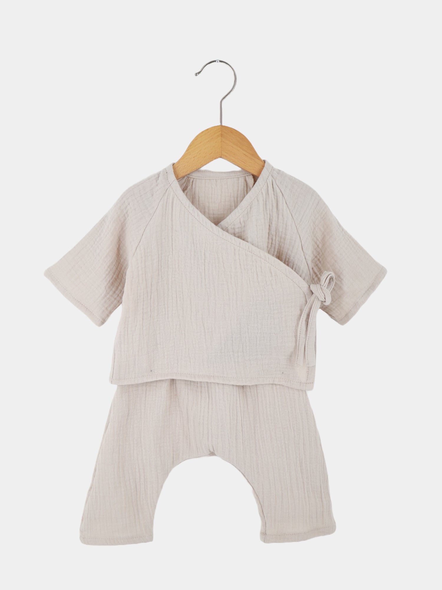 Babyset Baumwoll-Musselin - Sand: Ein weißes Wickel-Schlüttli aus atmungsaktivem Musselin, präsentiert auf einem Holzschwinger. Ideal für Neugeborene, mit seitlicher Schleifenbindung und weiten, krempelbaren Ärmeln.
