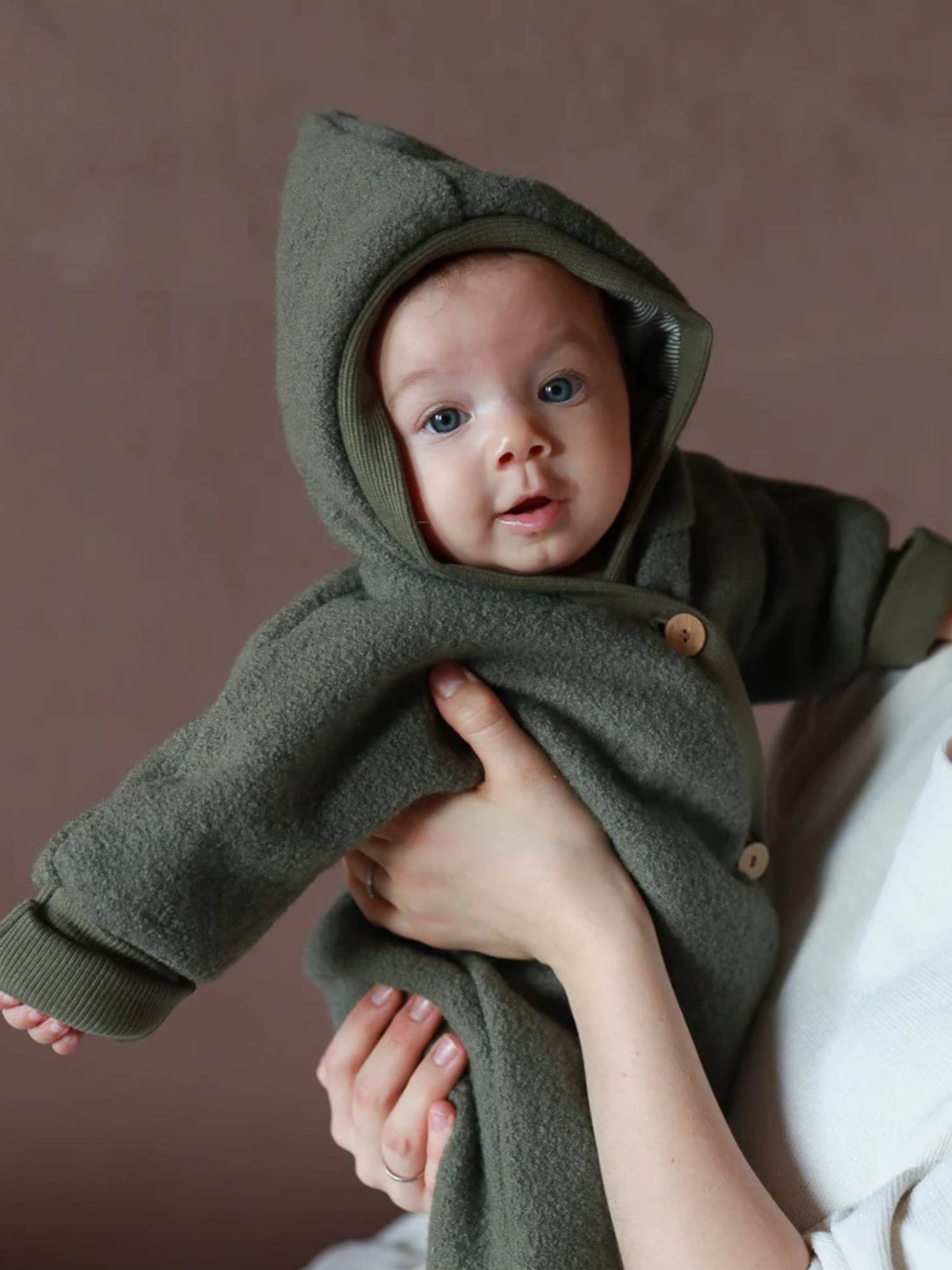 Ein Baby trägt den Kuschelanzug Wollwalk - Pistazie, einen warmen, olivgrünen, fleeceartigen Einteiler mit Kapuze, in den Armen eines Erwachsenen, vor einem schlichten Hintergrund.