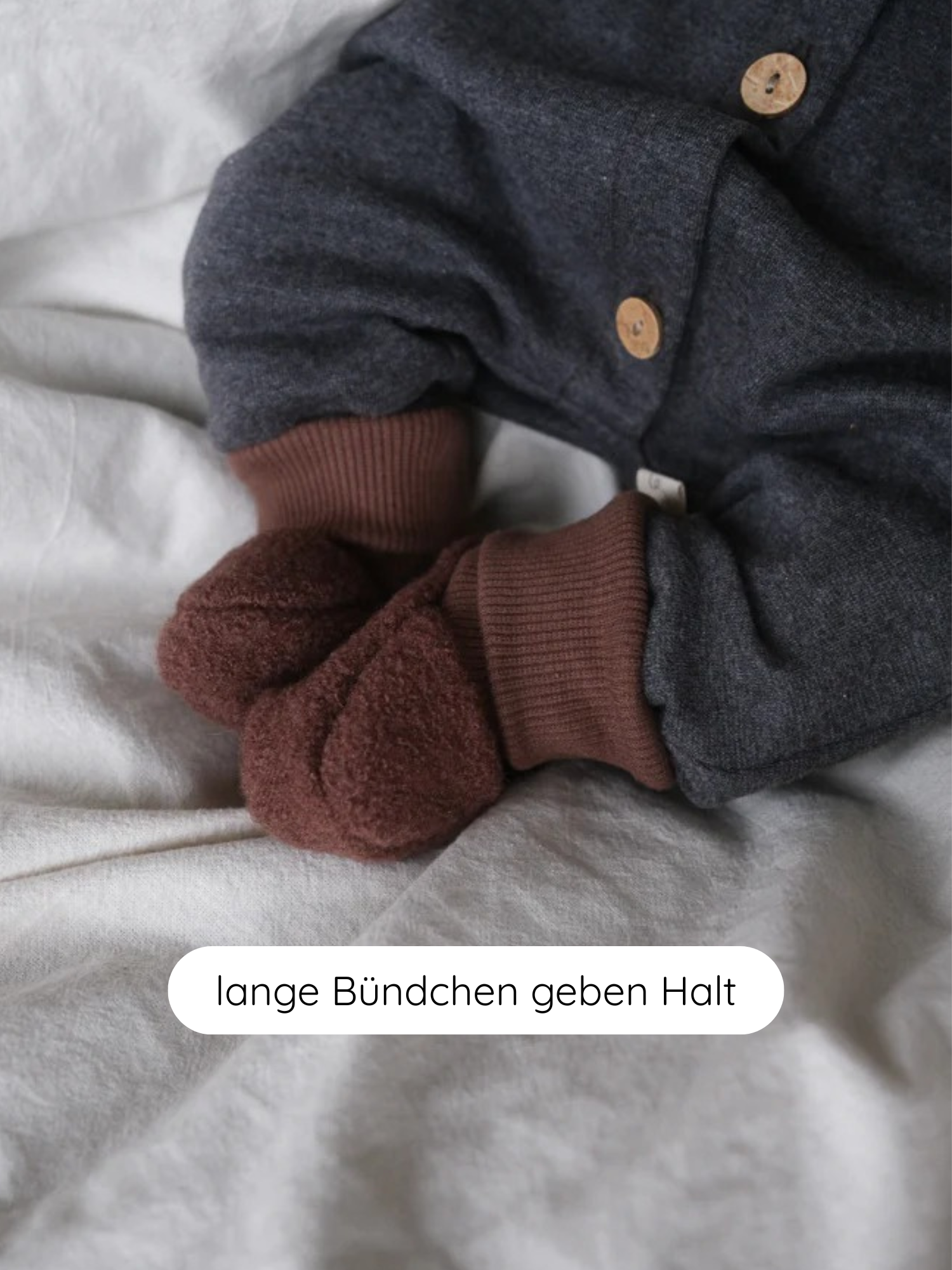 Babybooties Wollwalk - Schoko: Nahaufnahme zeigt ein Baby in einem dunkelgrauen Einteiler mit braunen, gerippten Bündchen, die in weiche, flauschige Booties übergehen, liegend auf einem hellen, zerknitterten Laken.