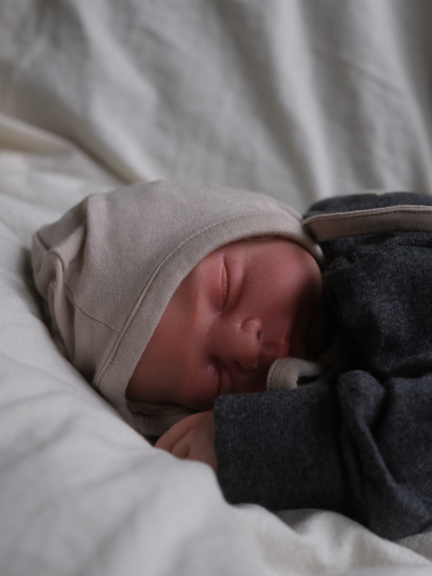 Schlafendes Baby mit dem Babyhäubchen Kaschmir-Mix - Sand auf weicher Unterlage, zeigt die kuschelige Passform und Elastizität der Mütze, passend zum Loopbody, für sanfte Wärme und Schutz.