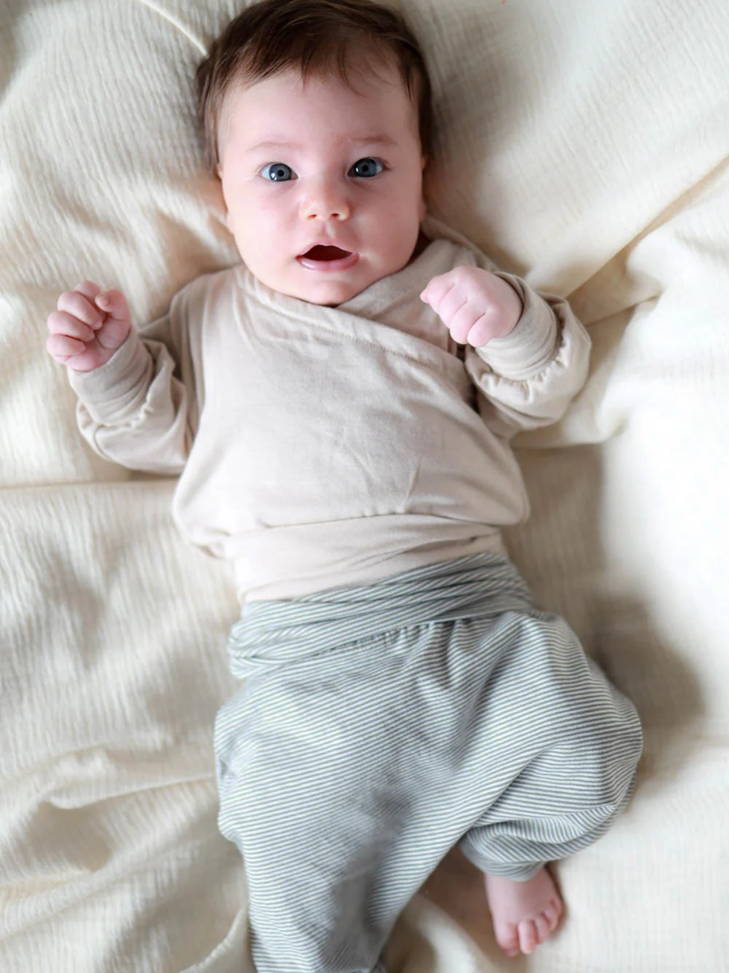 Baby in beiger Wickeljacke und Babyknicker Bio-Baumwolle - Maus Ringel, gestreifte grün-weiße Hose, auf Decke liegend, schaut mit leicht geballten Fäusten in die Kamera, bequem und nachhaltig.