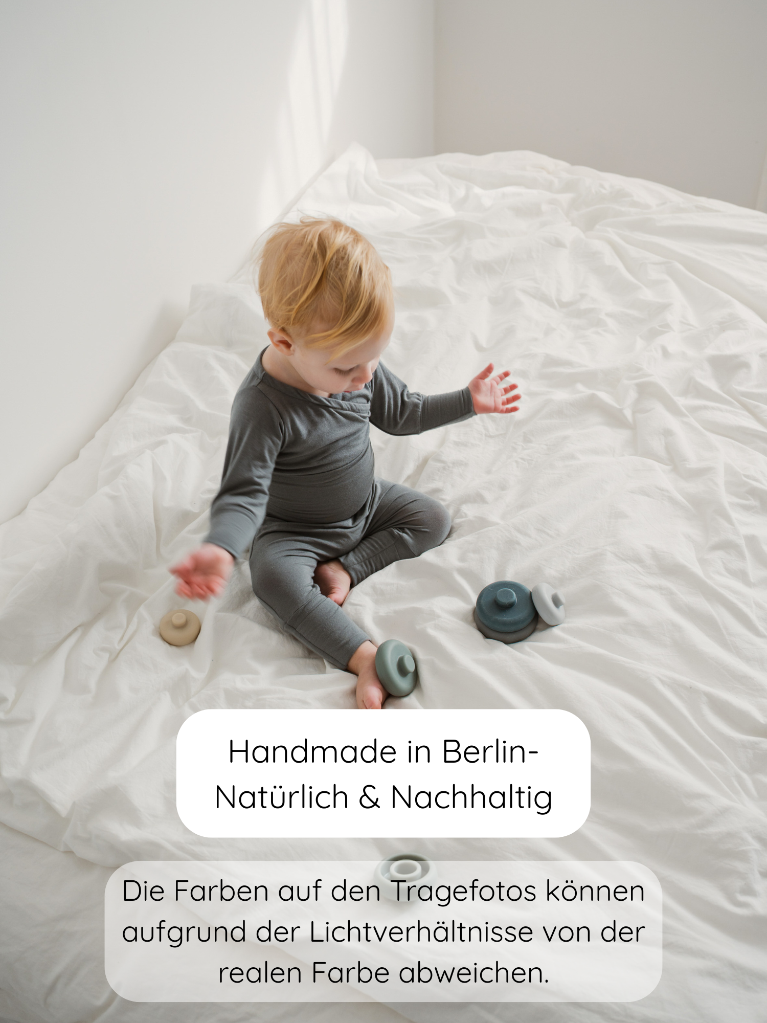 Kleines Kind in grauer Babyknicker Kaschmir-Mix - Taube auf weißen Laken, umgeben von natürlichen, handgefertigten Stapelspielzeugen. Text: Handmade in Berlin – Natürlich & Nachhaltig, mit Farbabweichungshinweis.