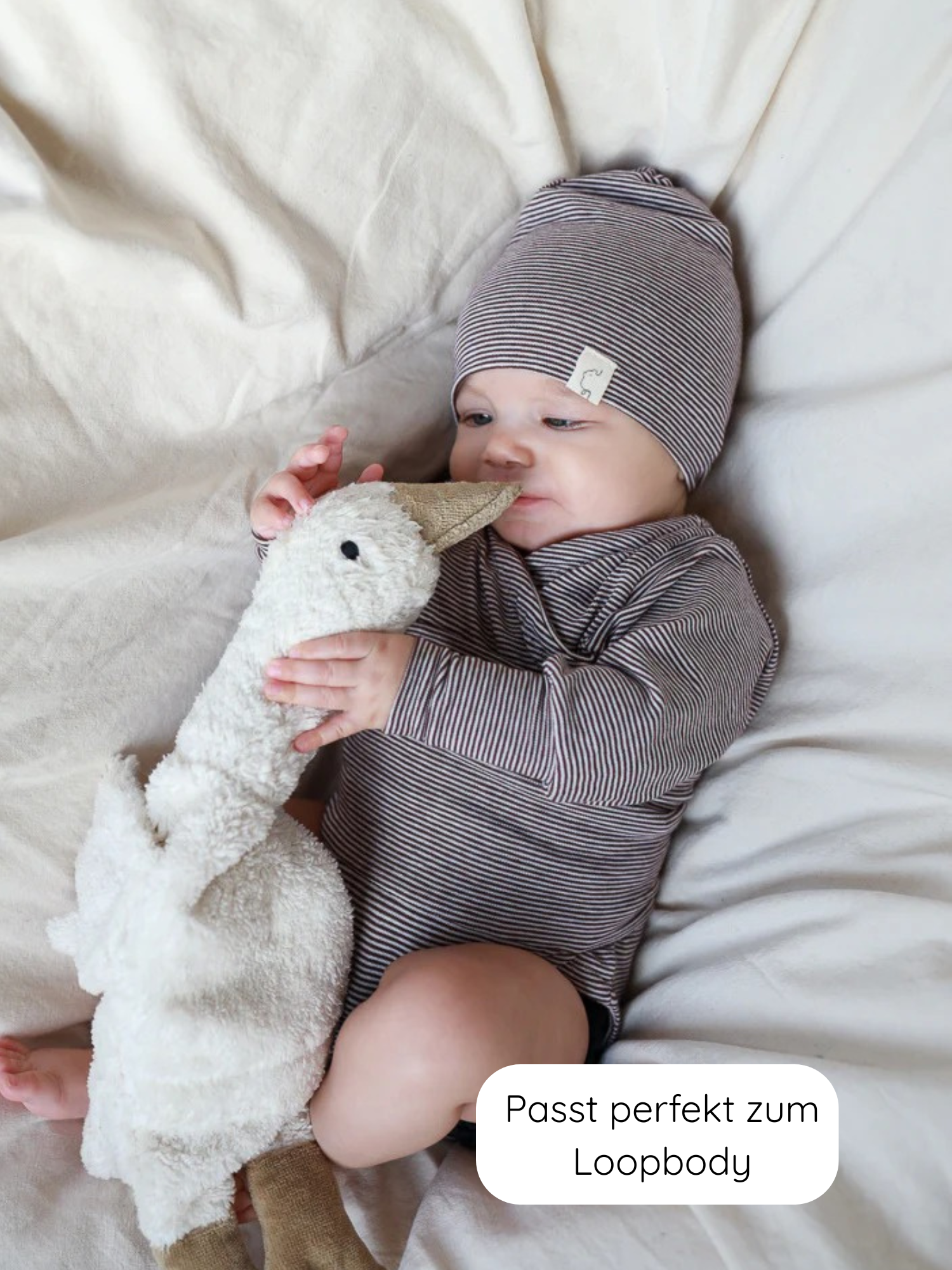 Baby in langärmligem, schmal gestreiftem Body und passender Mütze Beanie Bio-Baumwolle - Ringel Schoko aus 100% Bio-Baumwolle, kuschelt mit Plüschgans auf weicher, heller Unterlage.
