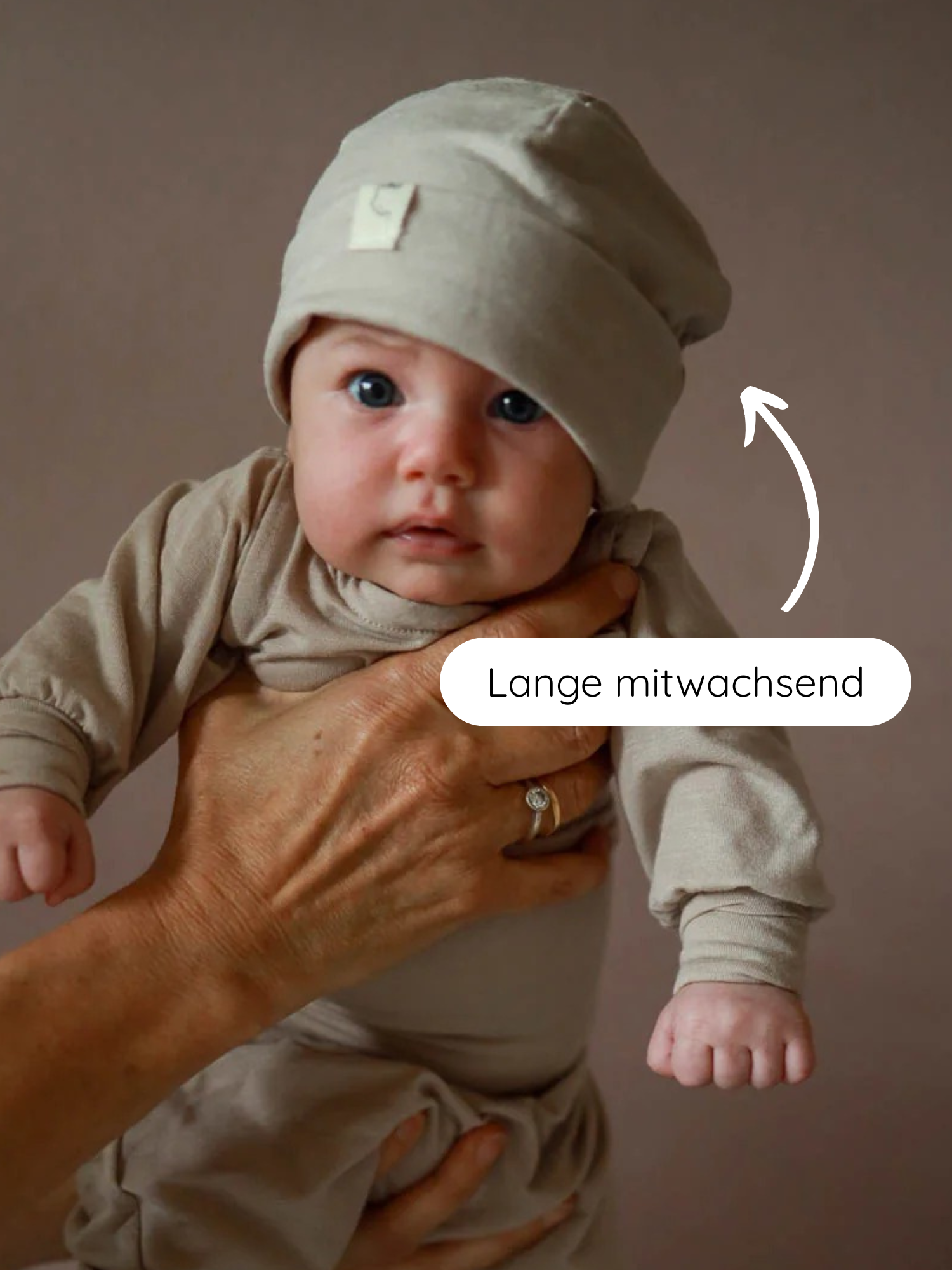 Baby in neutral-beiger Kleidung, gehalten von Erwachsenen. Beanie Kaschmir-Mix - Sand auf dem Kopf, mit Fokus auf die dehnbare, langlebige Mütze. Lange mitwachsend zeigt Anpassungsfähigkeit.