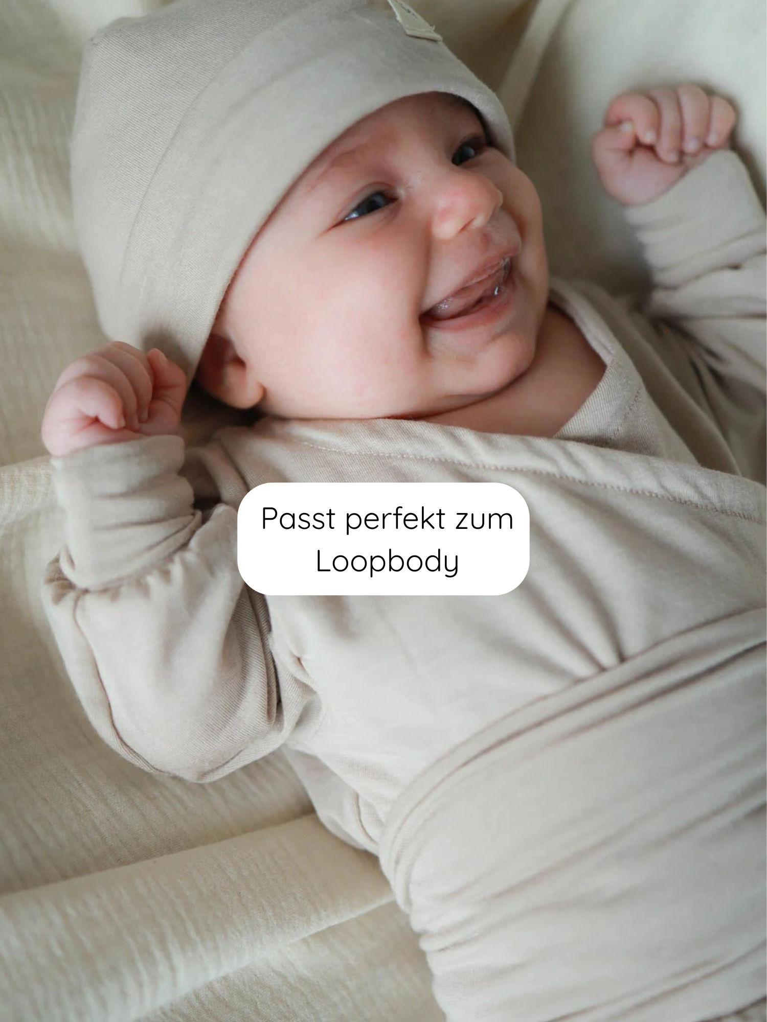 Lächelndes Baby trägt Beanie Kaschmir-Mix - Sand, kombiniert mit passendem Loopbody auf einer ähnlichen Decke, zeigt die weiche, dehnbare Mütze, die optimalen Komfort und klassischen Stil bietet.