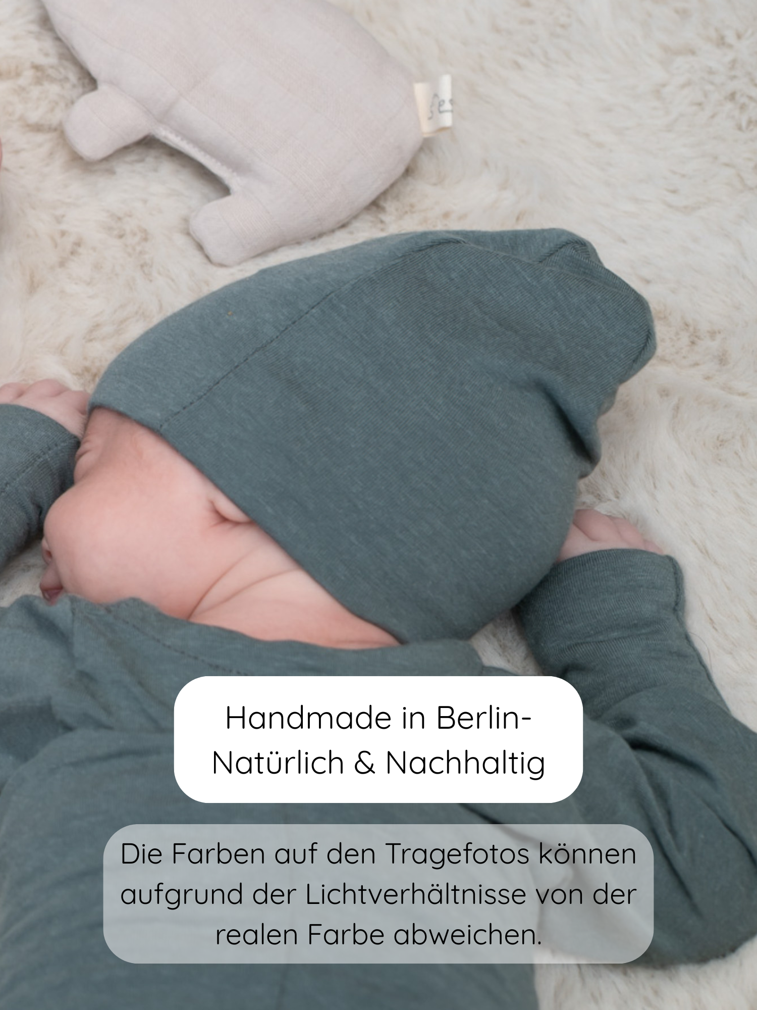 Baby auf cremefarbenem Teppich, trägt die Beanie Kaschmir-Mix - Taube in dezentem Grün, mit passendem Wickelanzug, neben einem beigen Kuscheltier. Text: Handmade in Berlin – Natürlich & Nachhaltig.