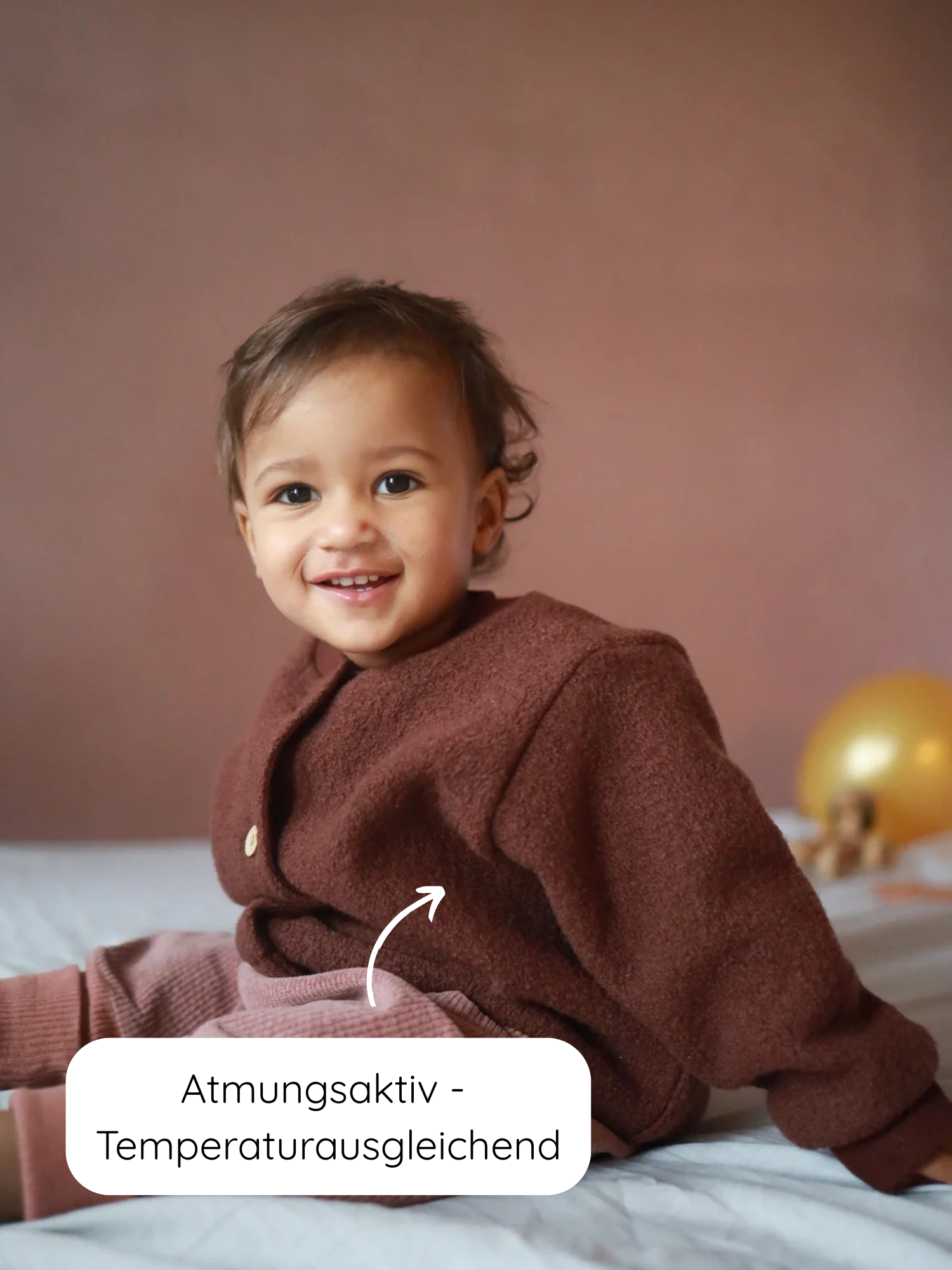 Kleinkind trägt eine Bomberjacke Wollwalk - Schoko mit strukturiertem Design, lächelt in die Kamera. Die Jacke bietet Funktionalität und Komfort, ideal für kühlere Jahreszeiten, mit atmungsaktiven, temperaturausgleichenden Eigenschaften.