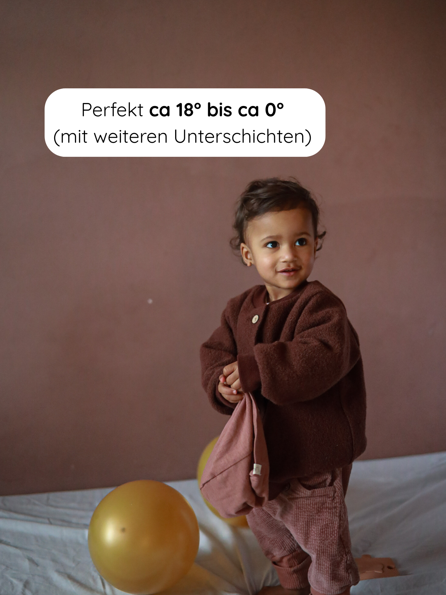 Kleinkind in Bomberjacke Wollwalk - Schoko, steht auf weißem Tuch, hält rosa Mütze. Goldener Ballon daneben. Jacke mit Reißverschluss, Taschen und Bündchen, ideal für kalte Temperaturen.