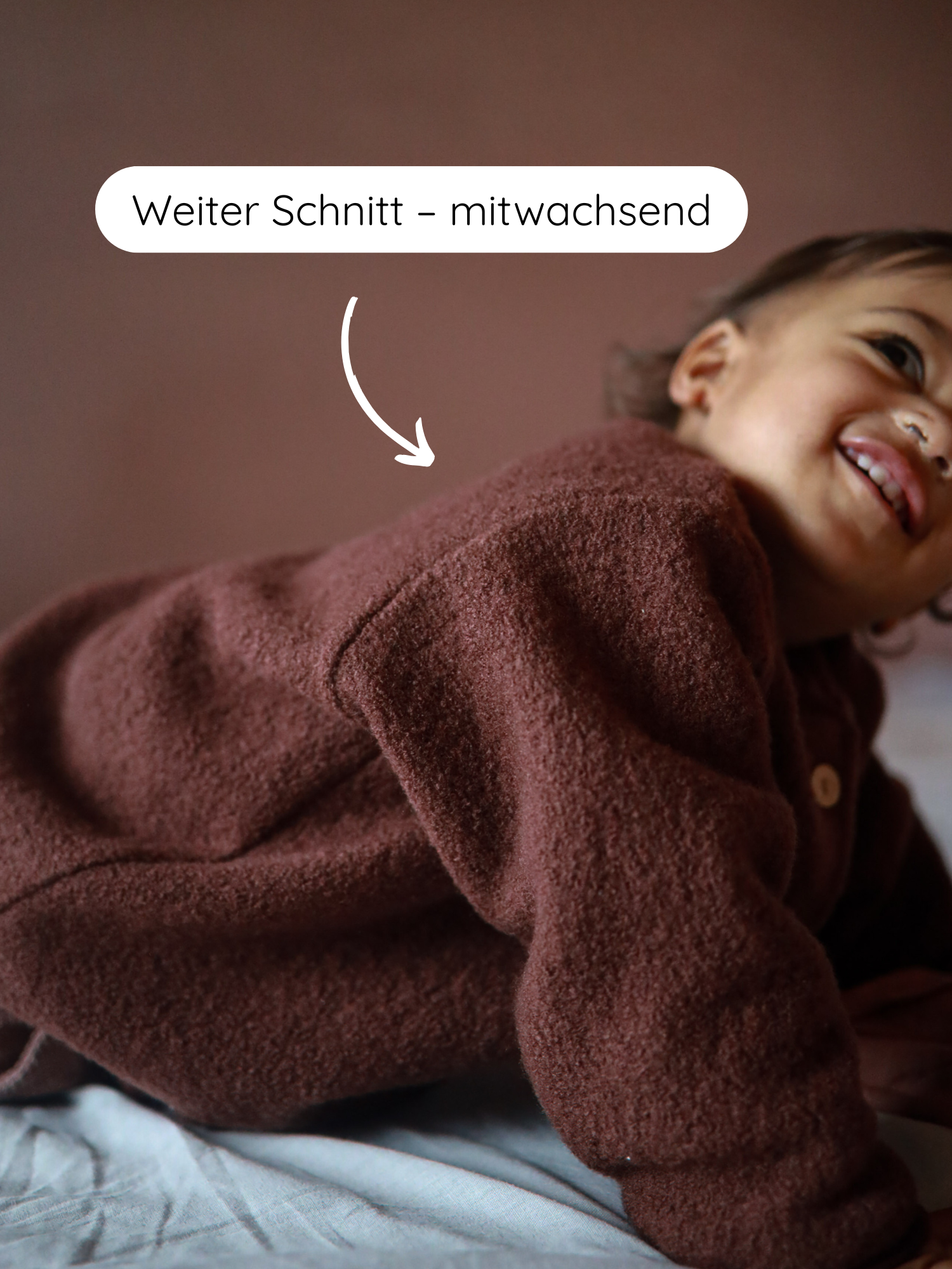 Baby in braunem Fleece-Einteiler, lächelnd, auf einem Bett. Sprechblase: „Weiter Schnitt – mitwachsend“. Produkt: Bomberjacke Wollwalk - Schoko mit durchgehendem Reißverschluss und elastischen Bündchen.