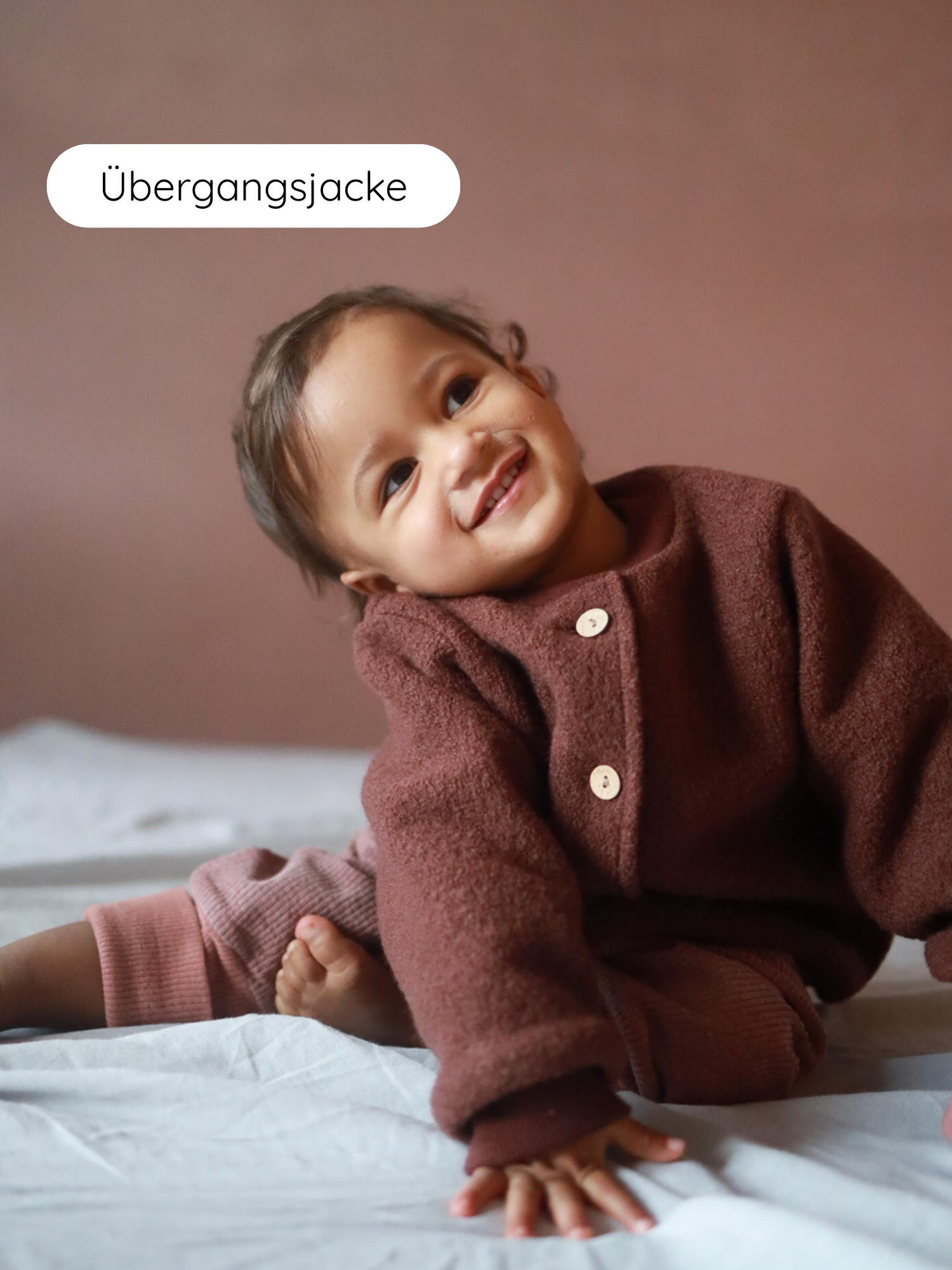 Kleinkind sitzt mit Bomberjacke Wollwalk - Schoko, einer rustbraunen Übergangsjacke mit Knöpfen, lächelnd auf einem weichen Untergrund. Die Jacke hat elastische Bündchen und einen modernen, kastigen Schnitt.