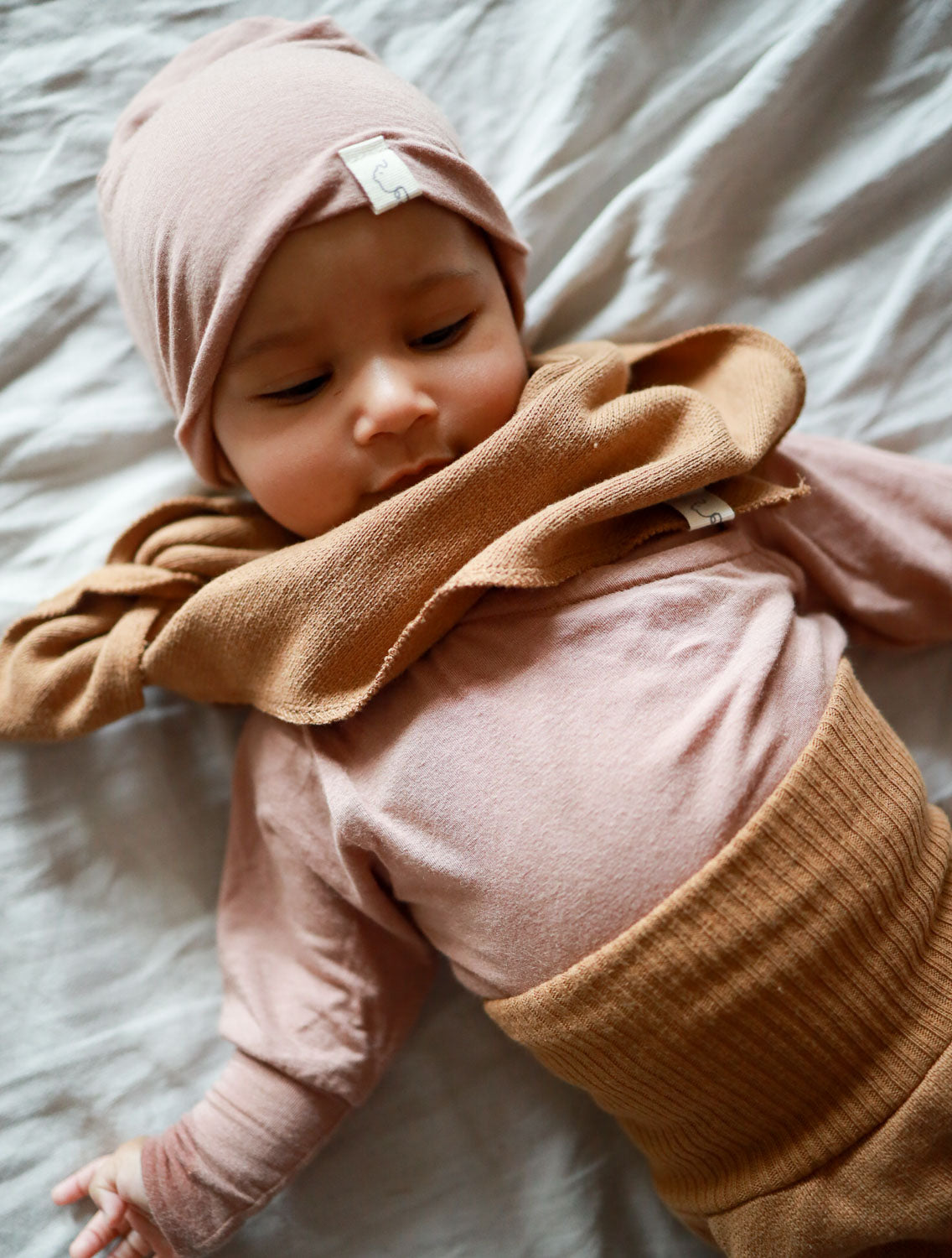 Baby auf Bett mit Dreieckstuch Strick - Sahara aus 100% recycelter Baumwolle, hält den Hals warm und kombiniert Stil mit Funktionalität.