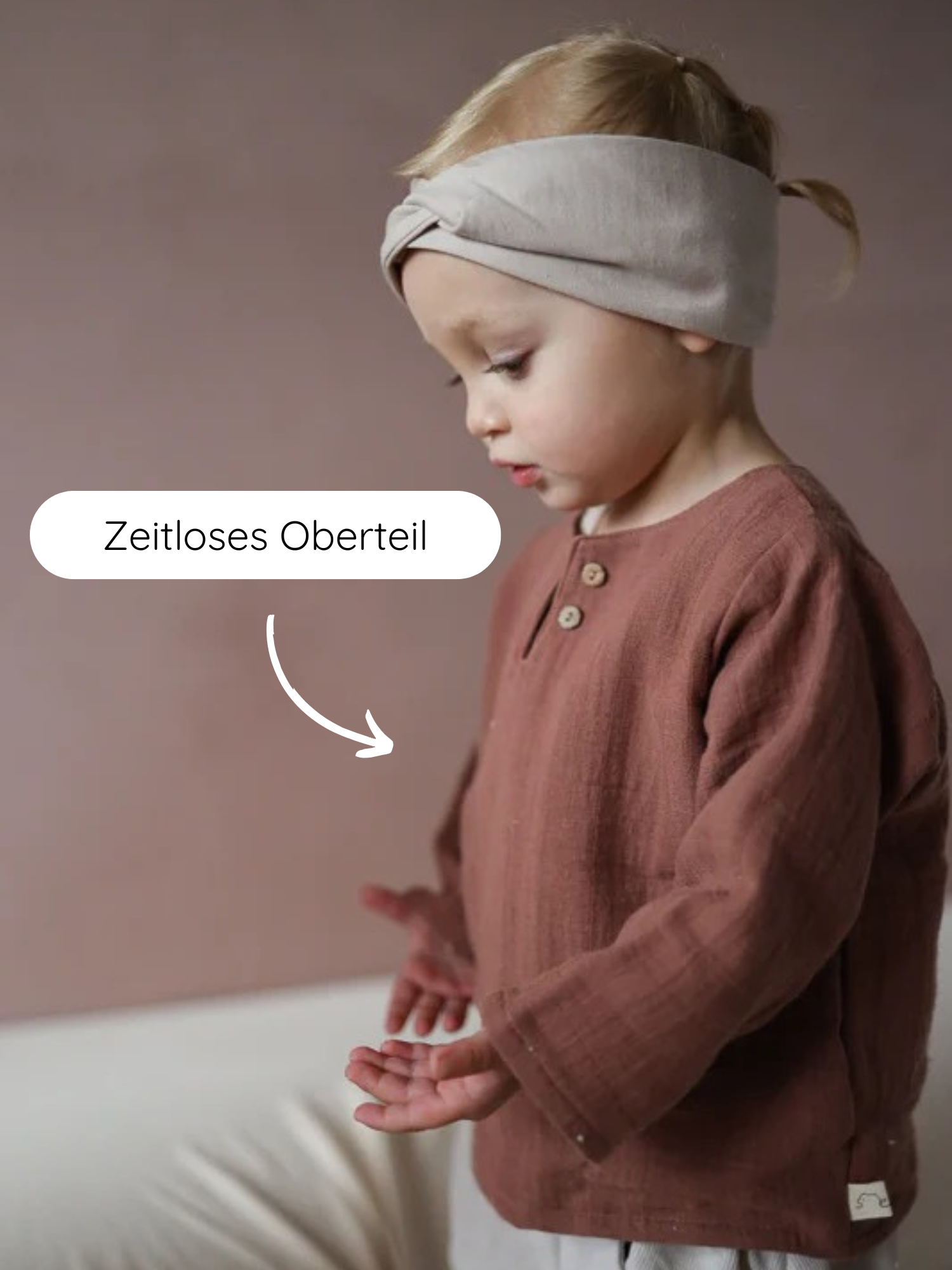 Kleinkind seitlich mit Hemdchen Bio-Musselin - Zimt, zeigt staubrosafarbenes Langarmoberteil mit Henley-Knopfleiste, kombiniert mit breitem, neutralem Stirnband. Zeitloses Oberteil als Overlay-Text hervorgehoben.