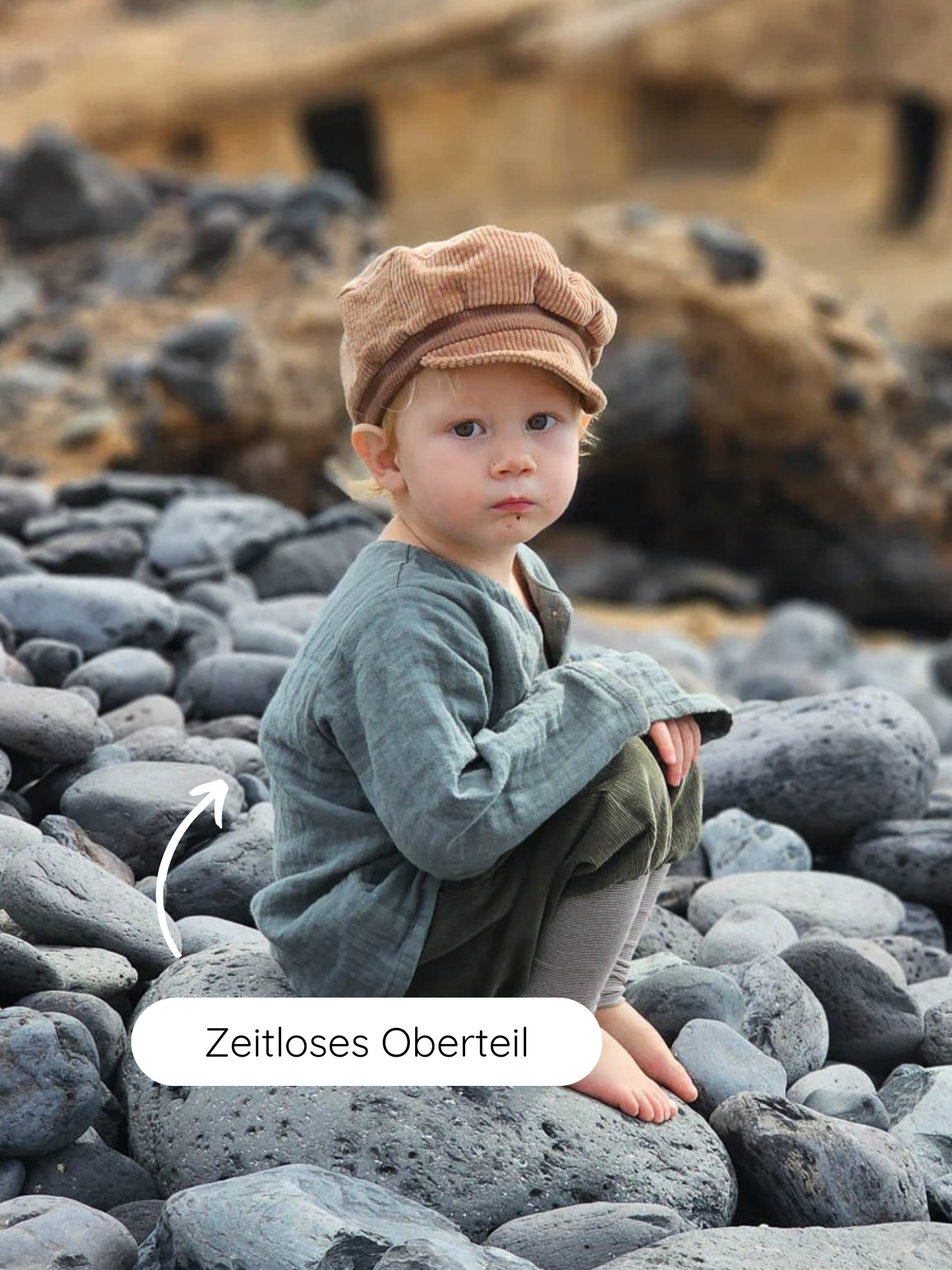 Kleinkind im Hemdchen Musselin - Eisblau sitzt barfuß auf dunklen Steinen. Das locker geschnittene, atmungsaktive Oberteil aus GOTS-zertifiziertem Bio-Musselin ist perfekt für warme Tage.