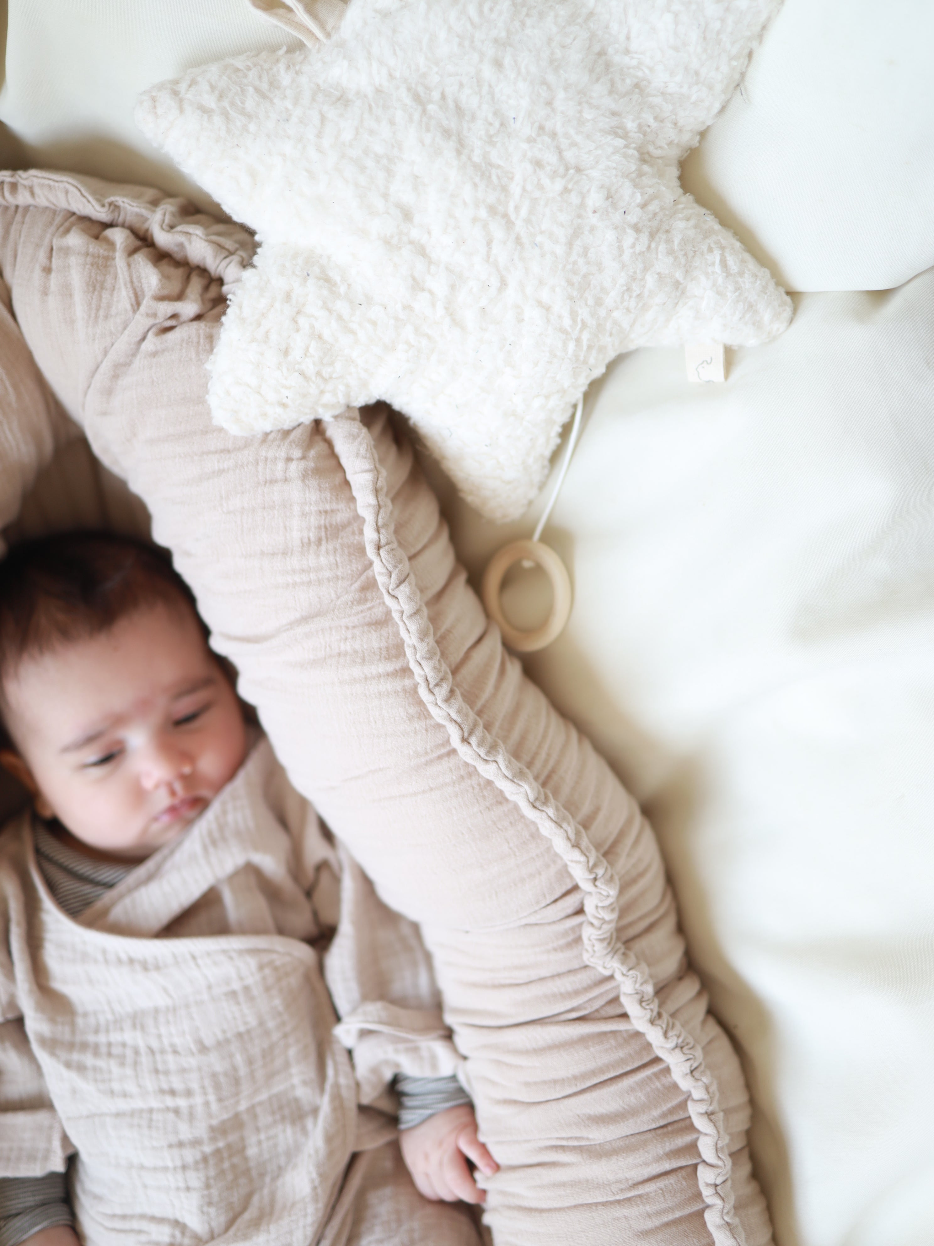 Ein Baby liegt friedlich in einem beigen Nest, eingehüllt in das Baby-Set Baumwoll-Musselin - Sand mit einem Wickel-Schlüttli und weichen Hosen aus atmungsaktivem Musselin, daneben ein Sternkissen.