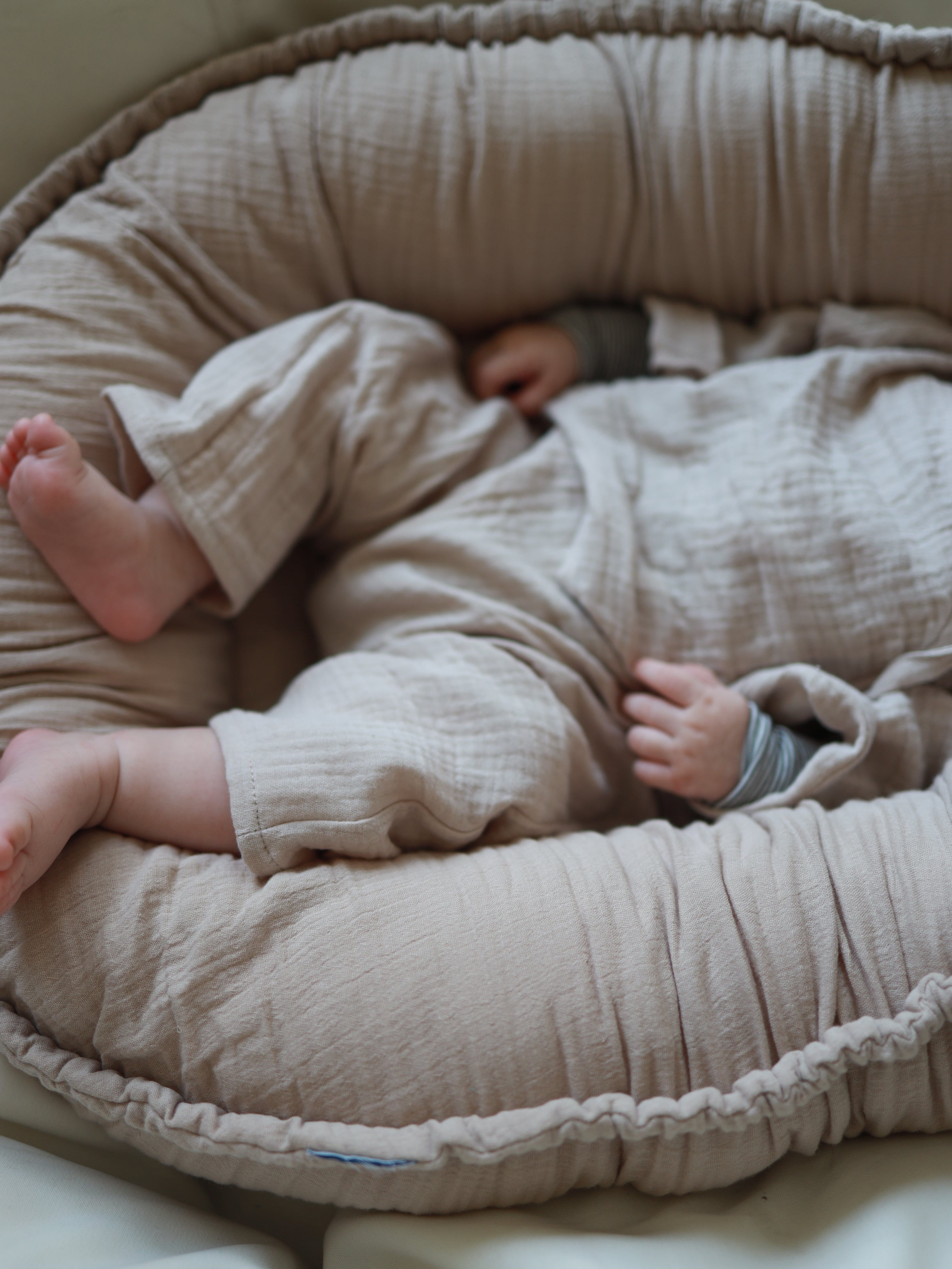Ein Baby liegt im Baby-Set Baumwoll-Musselin - Sand, bestehend aus Wickel-Schlüttli und Hose, bequem im Baby-Nest. Kleidung und Nest sind farblich abgestimmt, aus weichem Musselin gefertigt.