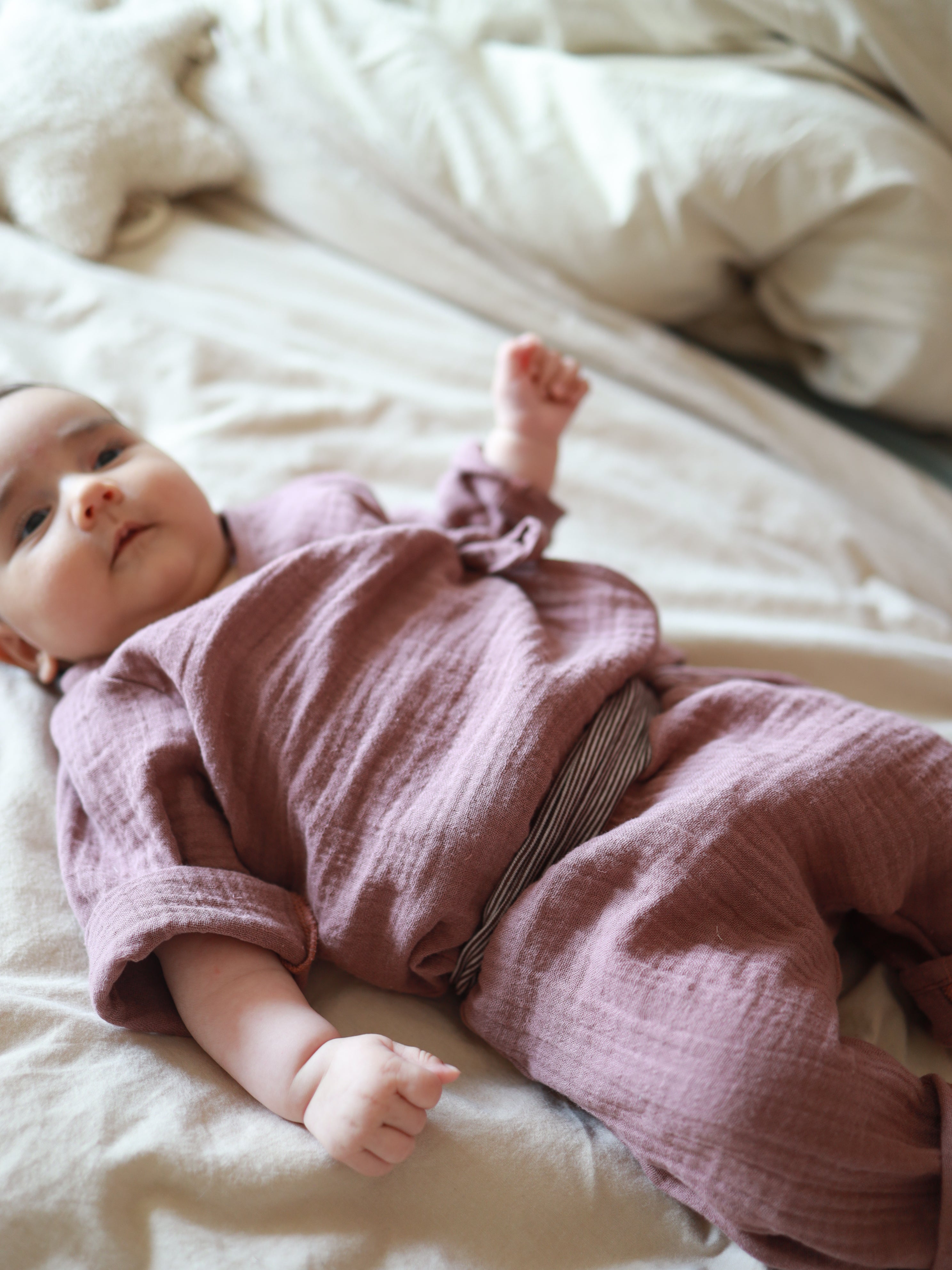 Baby-Set Baumwoll-Musselin - Malve: Ein Baby liegt auf dem Rücken, trägt ein weiches Musselin-Wickel-Schlüttli und Hose. Im Hintergrund ein plüschiges, sternförmiges Kissen.