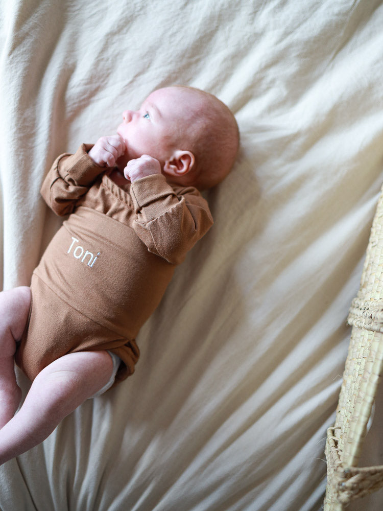 Neugeborenes in einem braunen, langärmeligen Onesie mit dem Namen Toni, veranschaulicht die Individuelle Stickerei. Rechts ist ein Teil eines geflochtenen Korbes sichtbar.